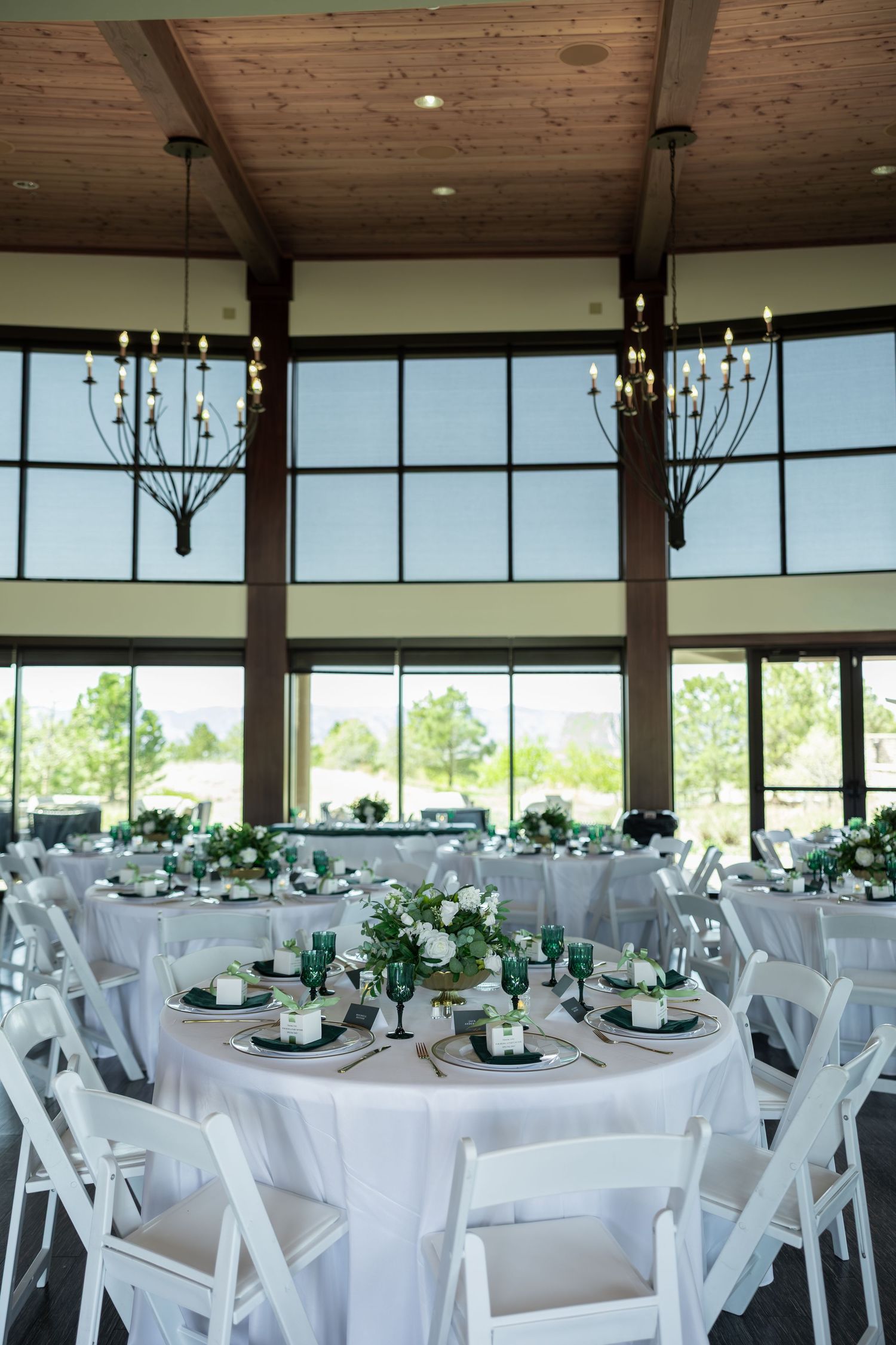 Bright wedding reception hall with white round tables, green glassware, floral centerpieces, and modern chandeliers.