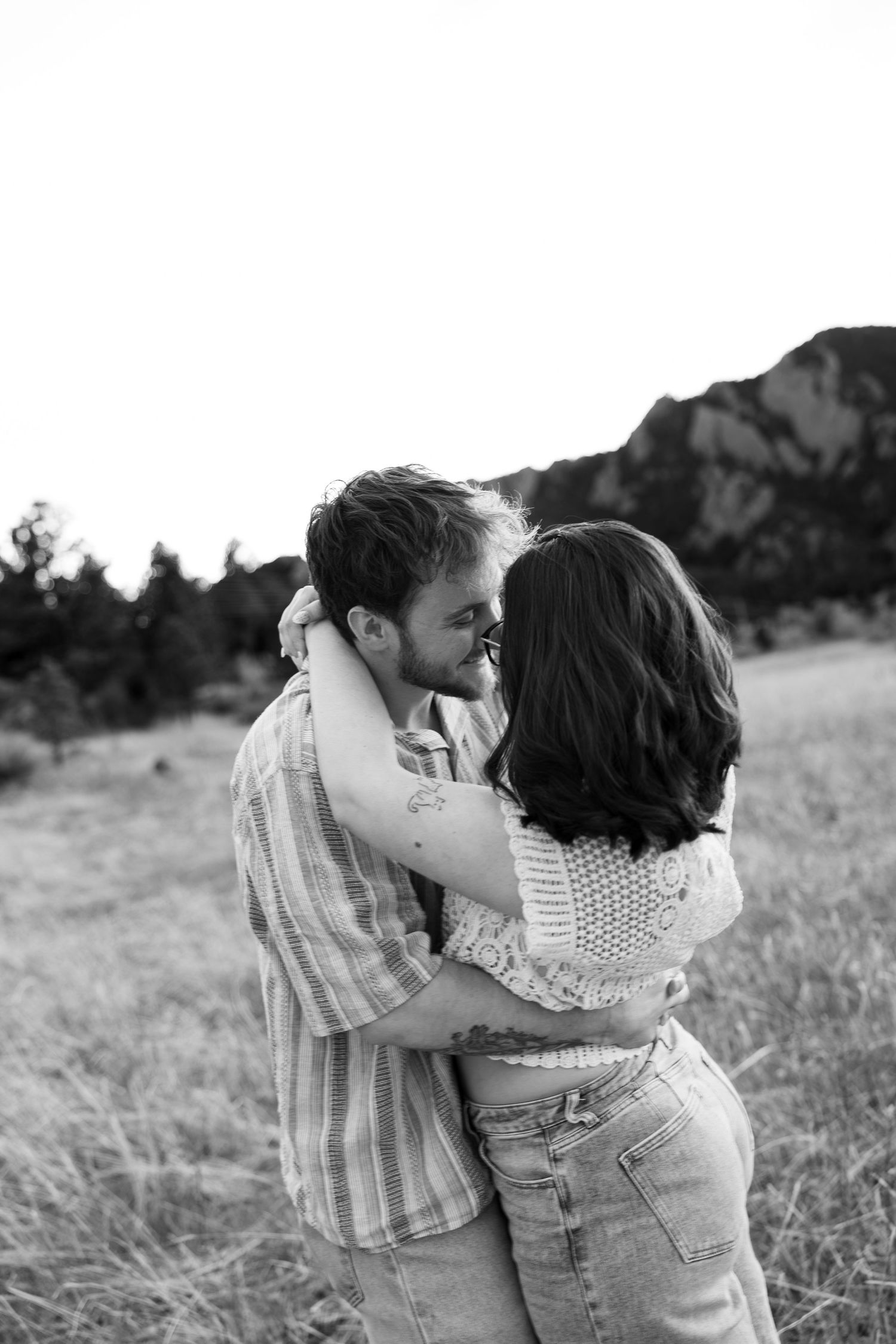 Black and white photo of a couple embracing and kissing in a field with rocky mountains in the background.