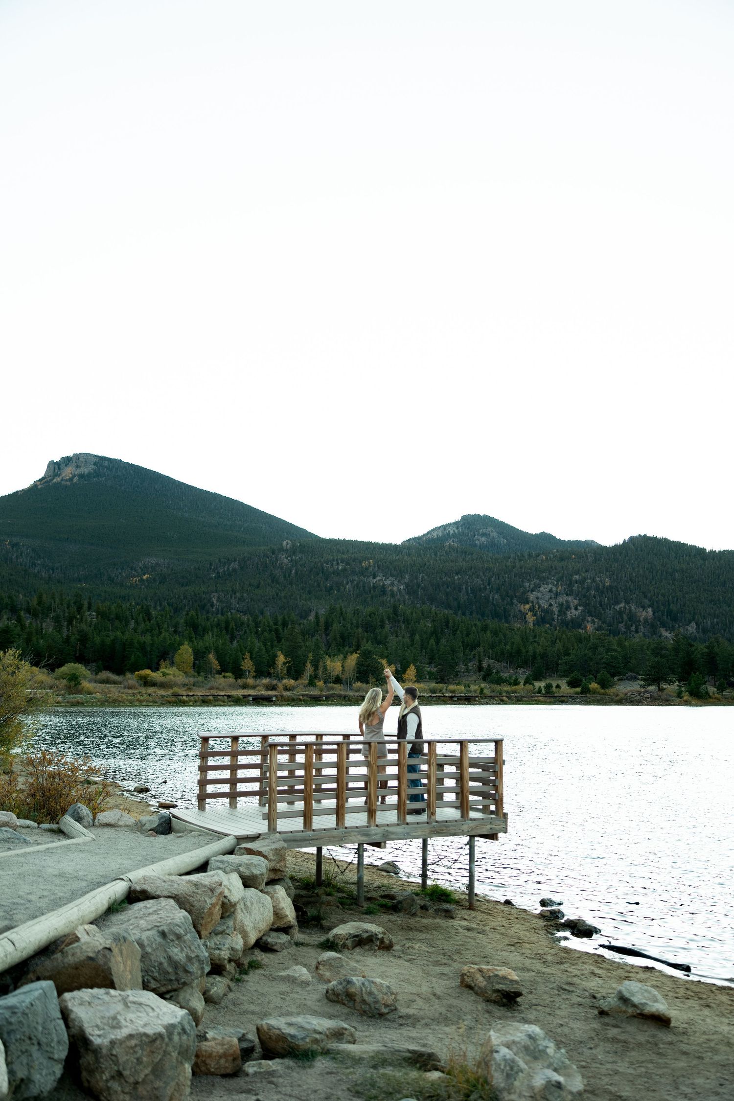 Couple stands on a wooden lakeside pier raising their arms, with rocky shoreline, calm water and forested mountains behind them.