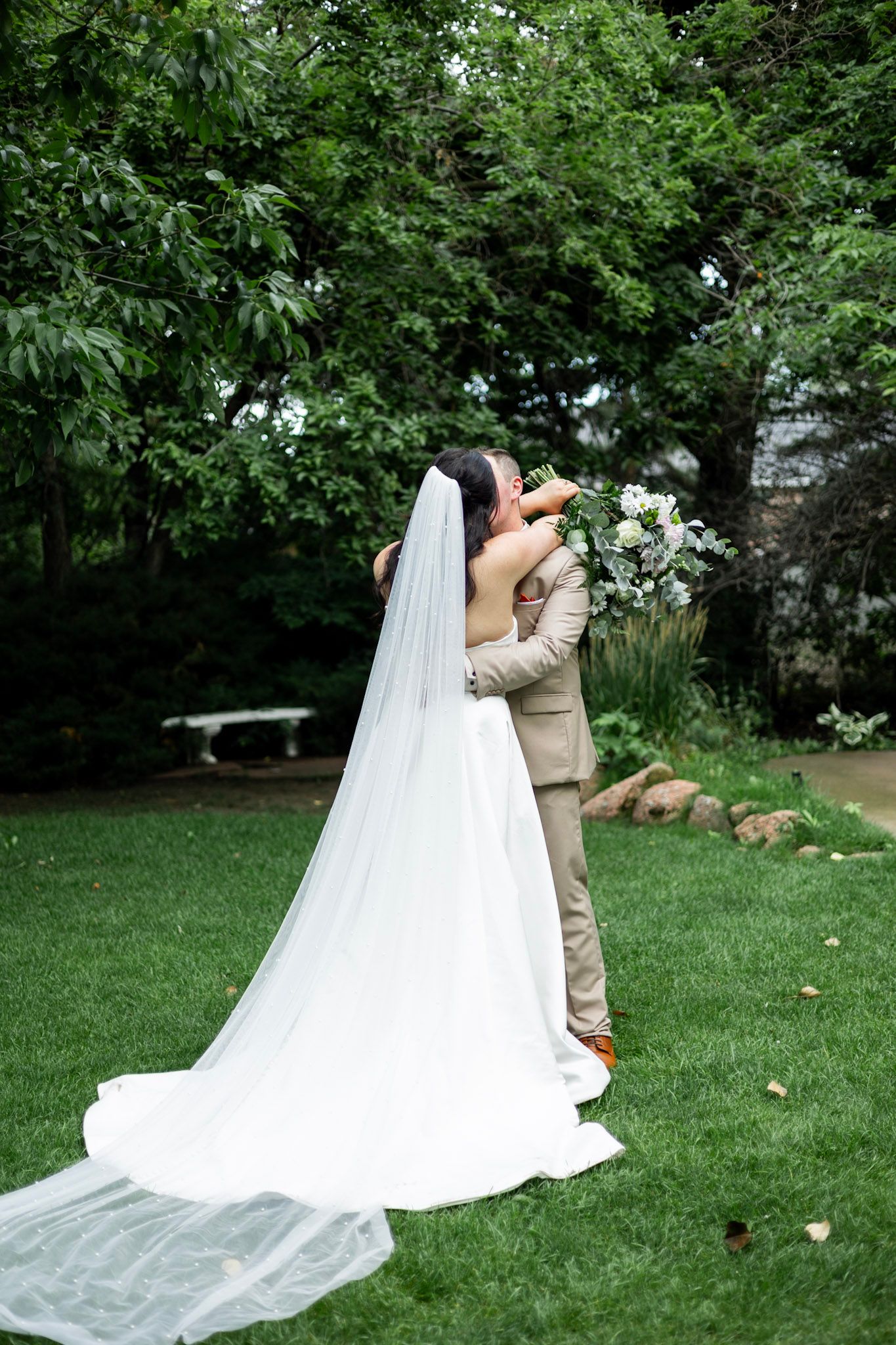 A bride and groom share an intimate embrace on a lush green lawn with flowing white veil trailing behind them.