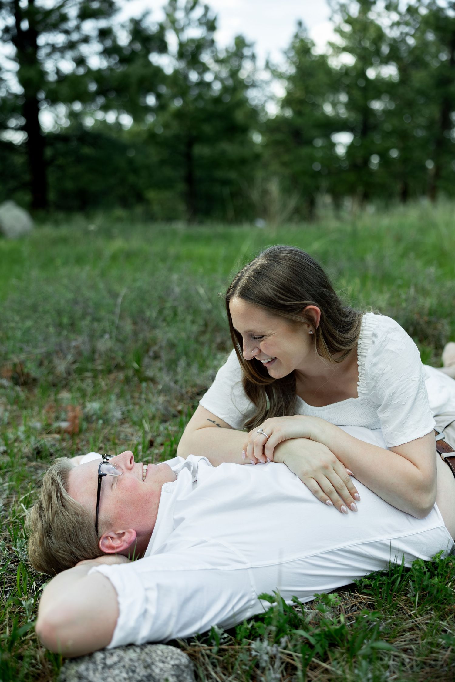 Couple lying in green grass, woman in white dress smiling at man in white shirt wearing glasses, outdoor romantic moment.