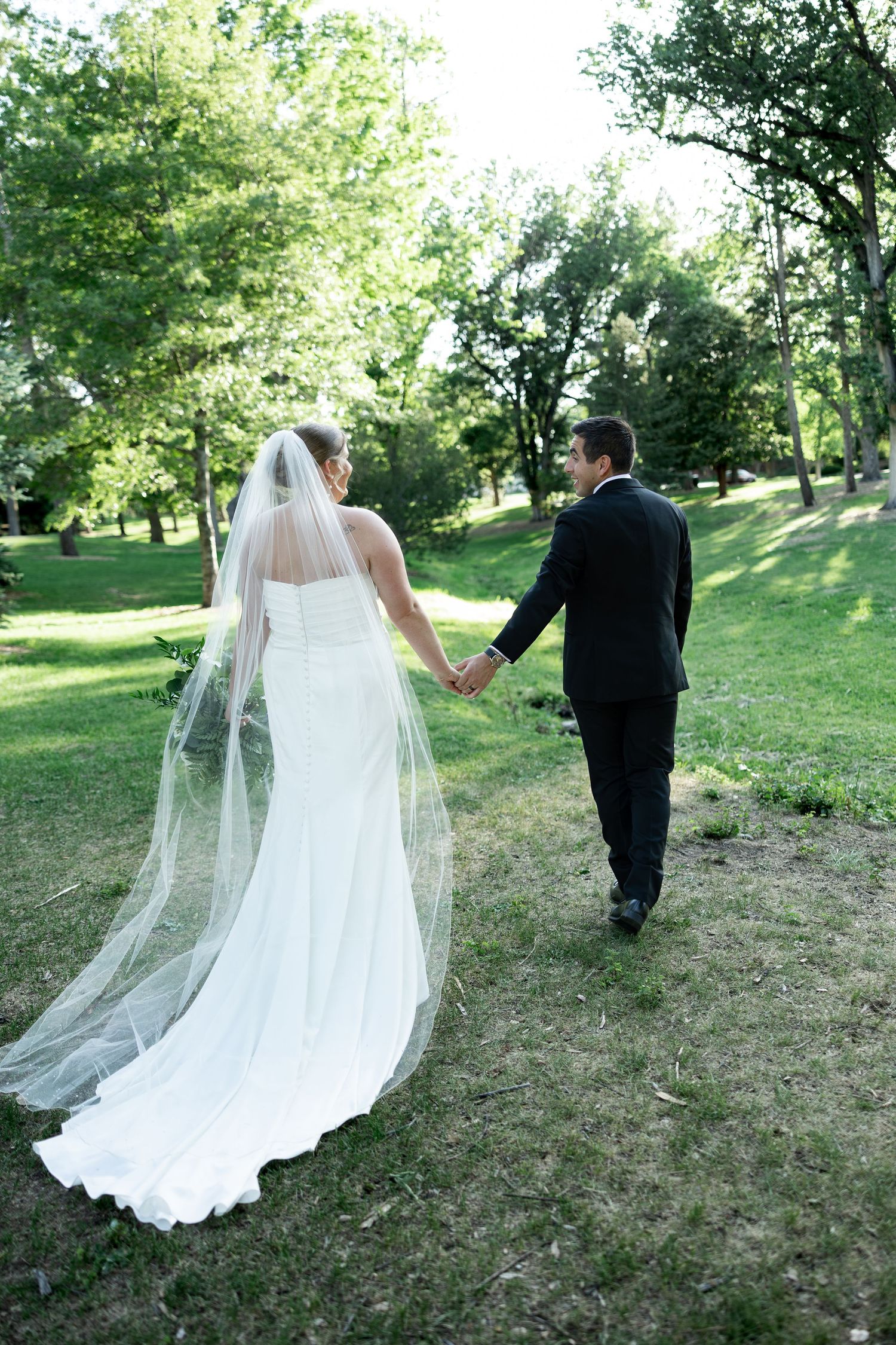 Bride and groom holding hands walking through lush green park, bride wearing white gown with long flowing veil.