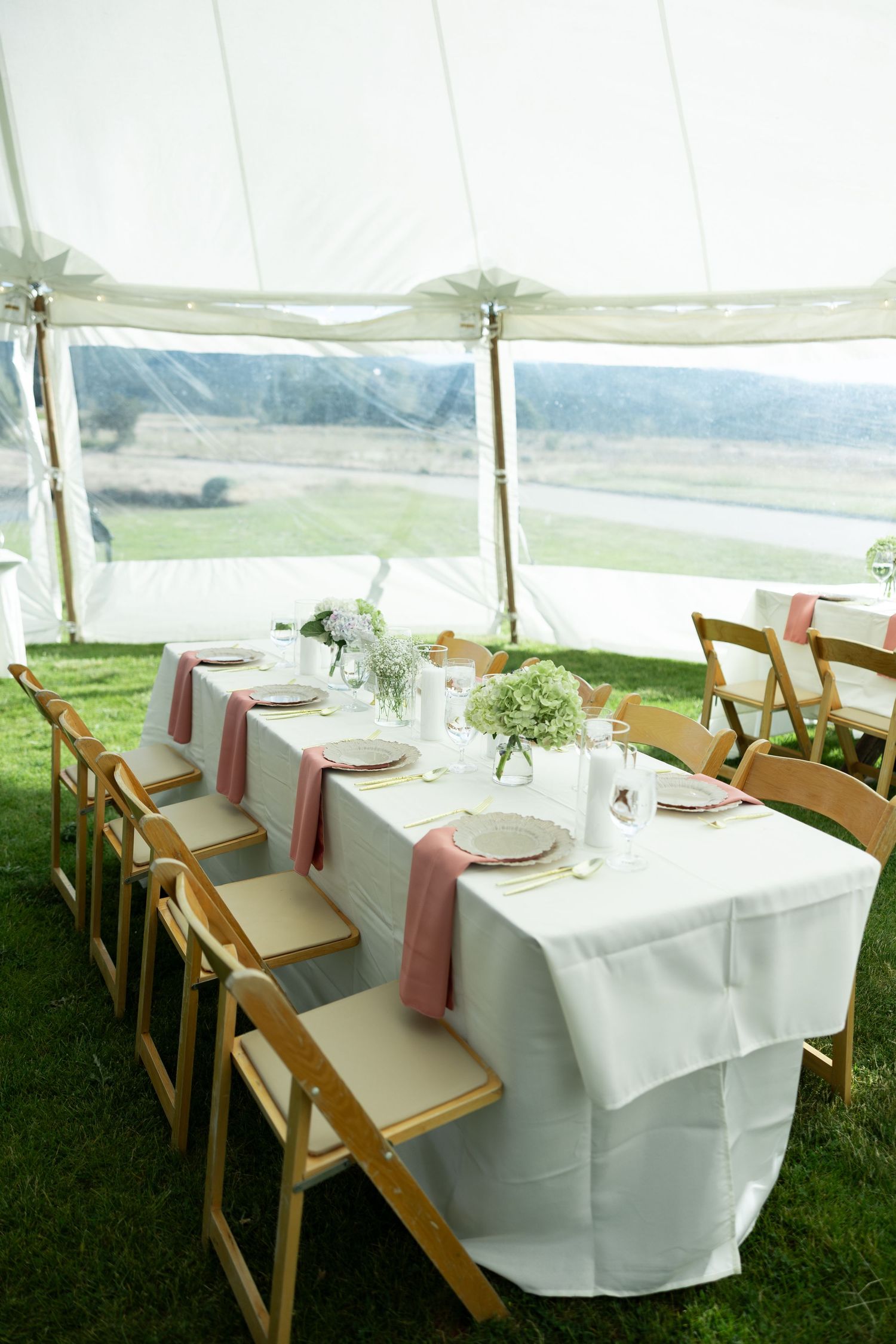 Elegant outdoor wedding reception table set under a white tent with pink napkins, floral centerpieces, and wooden folding chairs.