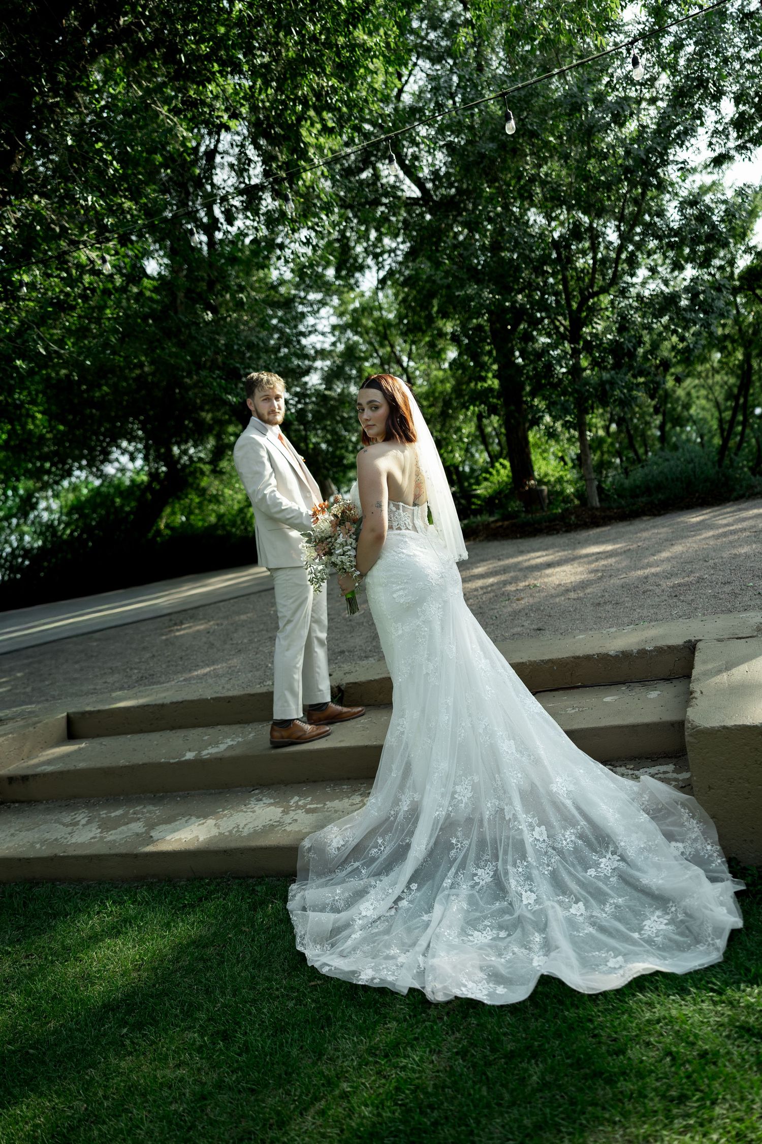 Bride in lace gown with train stands near groom in beige suit on garden steps surrounded by lush trees.