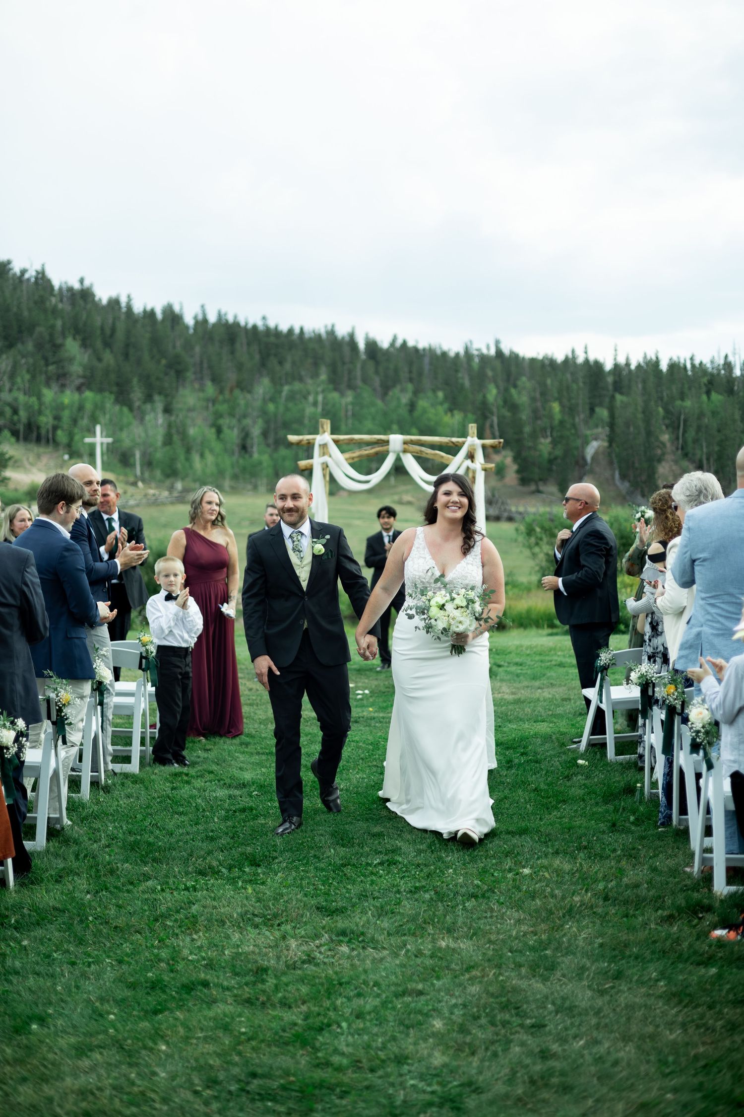 Wedding guests gather on a grassy hillside for an outdoor ceremony with white chairs and mountain views in the background.