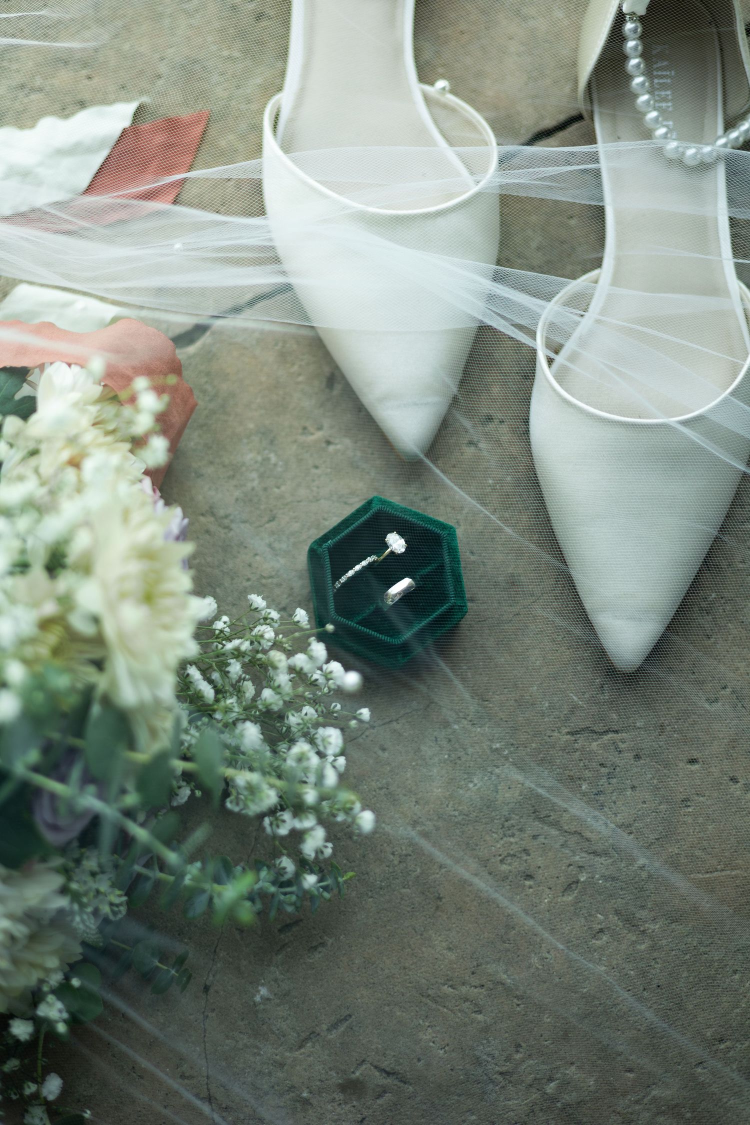 White bridal shoes and a wedding ring box displayed with delicate white flowers on a stone surface.