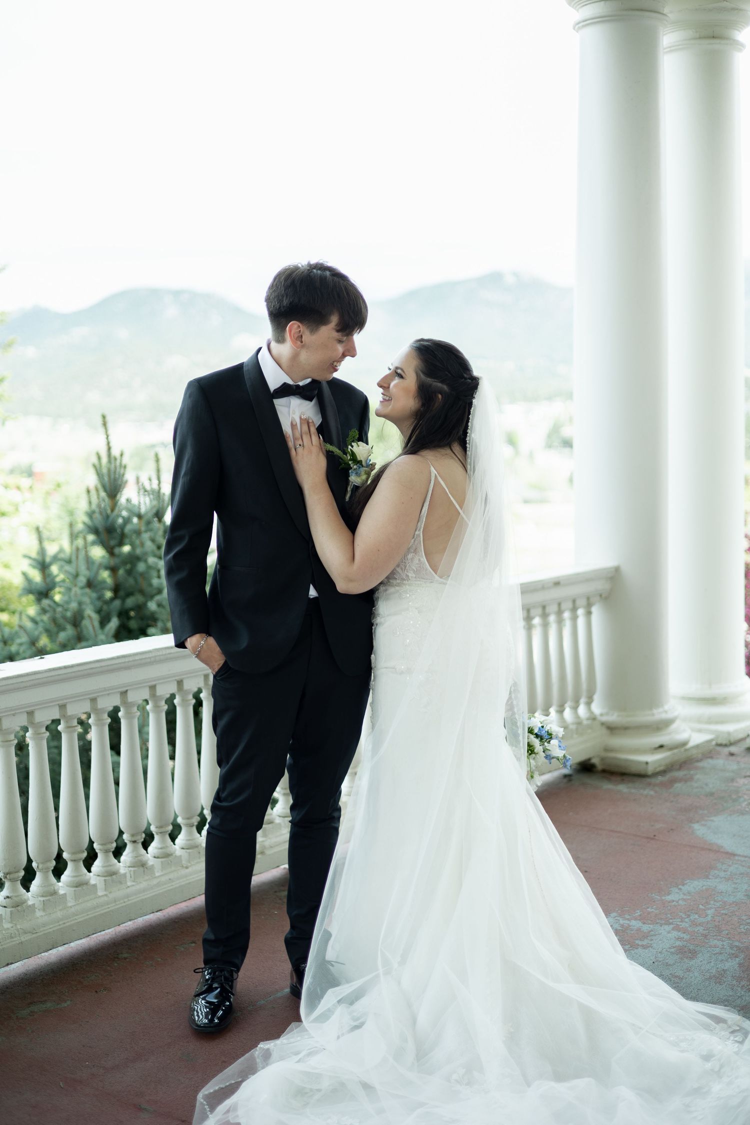 A wedding couple shares an intimate moment on an elegant balcony with white columns and mountain views in the background.