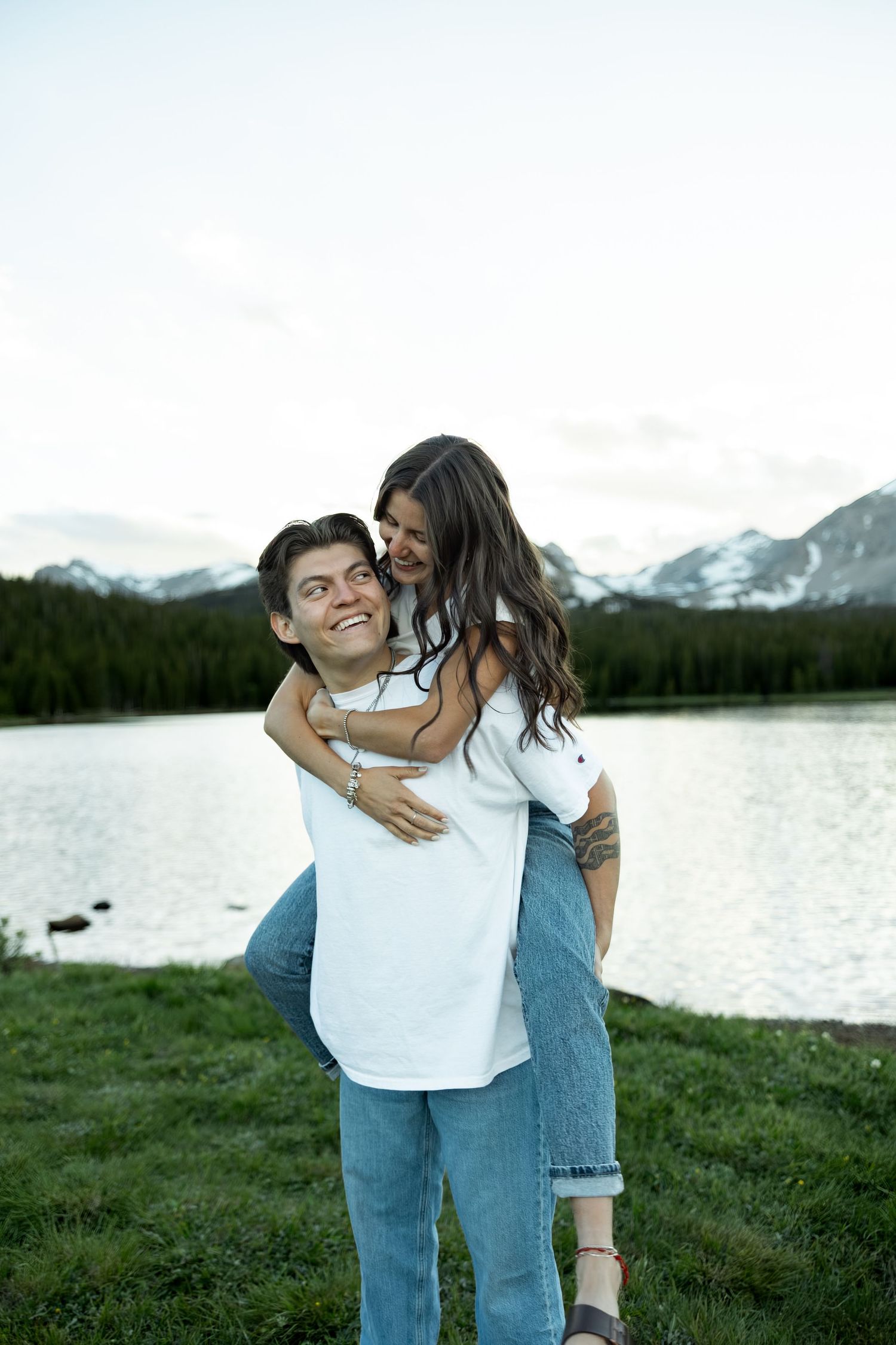 Happy couple sharing a piggyback ride by a serene mountain lake with snow-capped peaks and evergreen forest behind them.