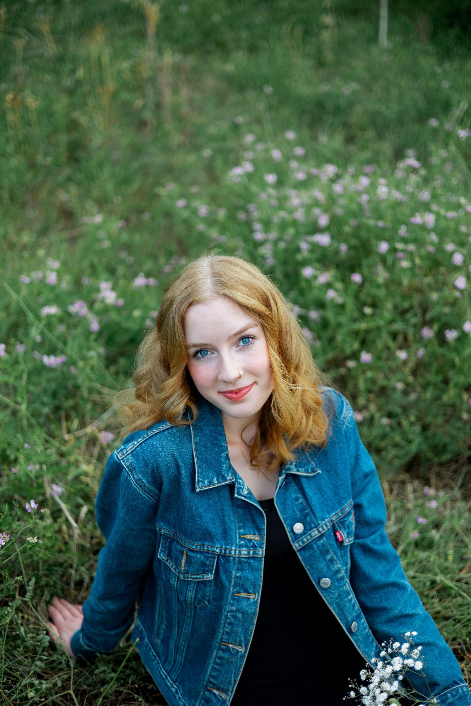 A person with red hair wearing a denim jacket poses in a field of wildflowers on a sunny day.