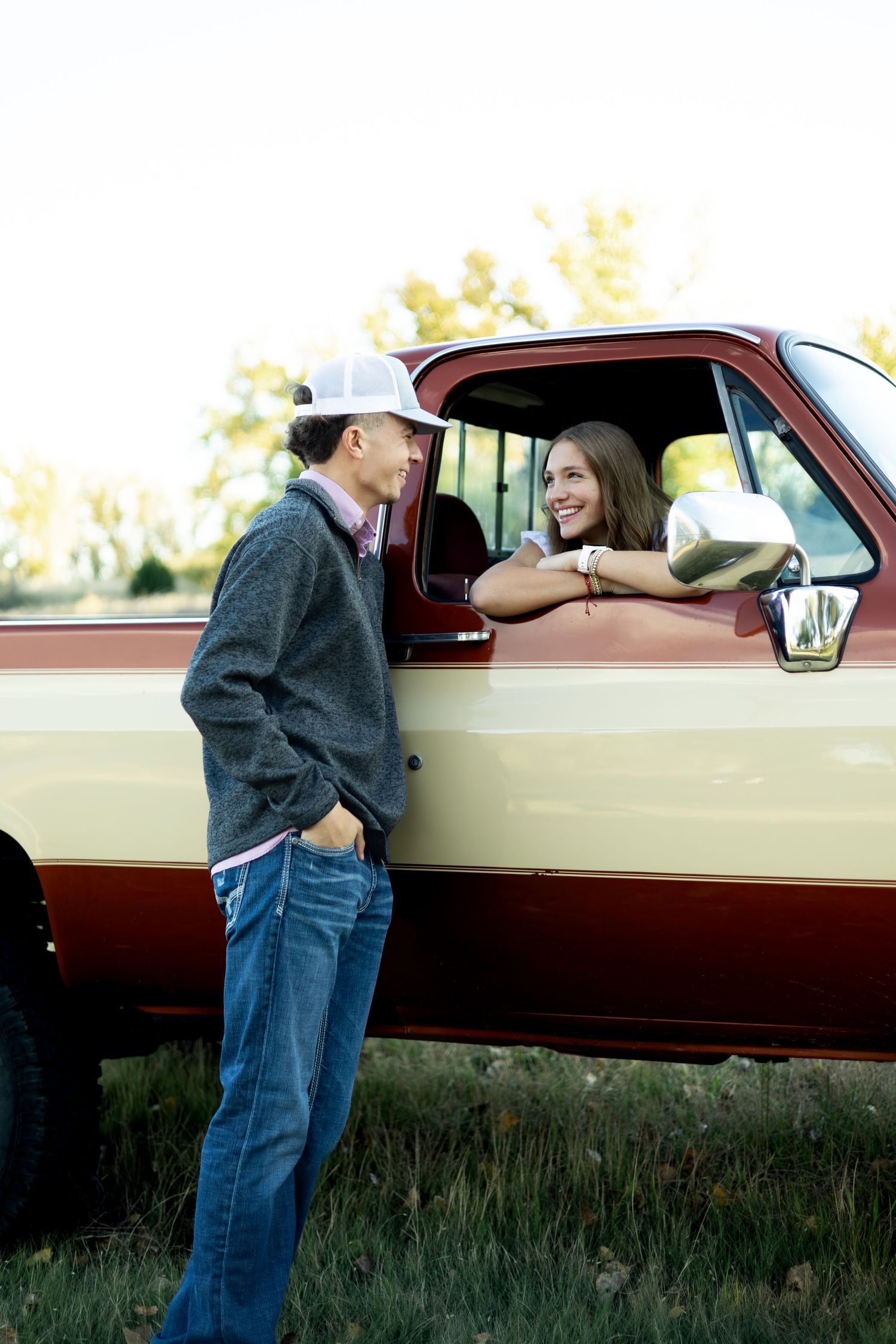 A person in jeans and jacket leans into the window of a vintage burgundy car parked in a grassy field at sunset.