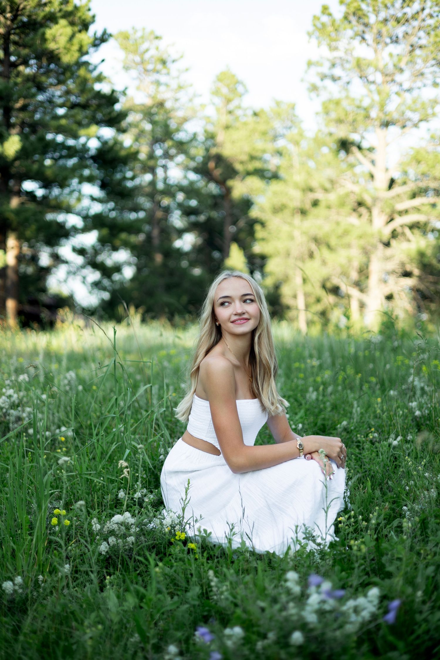 A young woman in a white dress sits gracefully among wildflowers and tall grass in a sunny meadow.