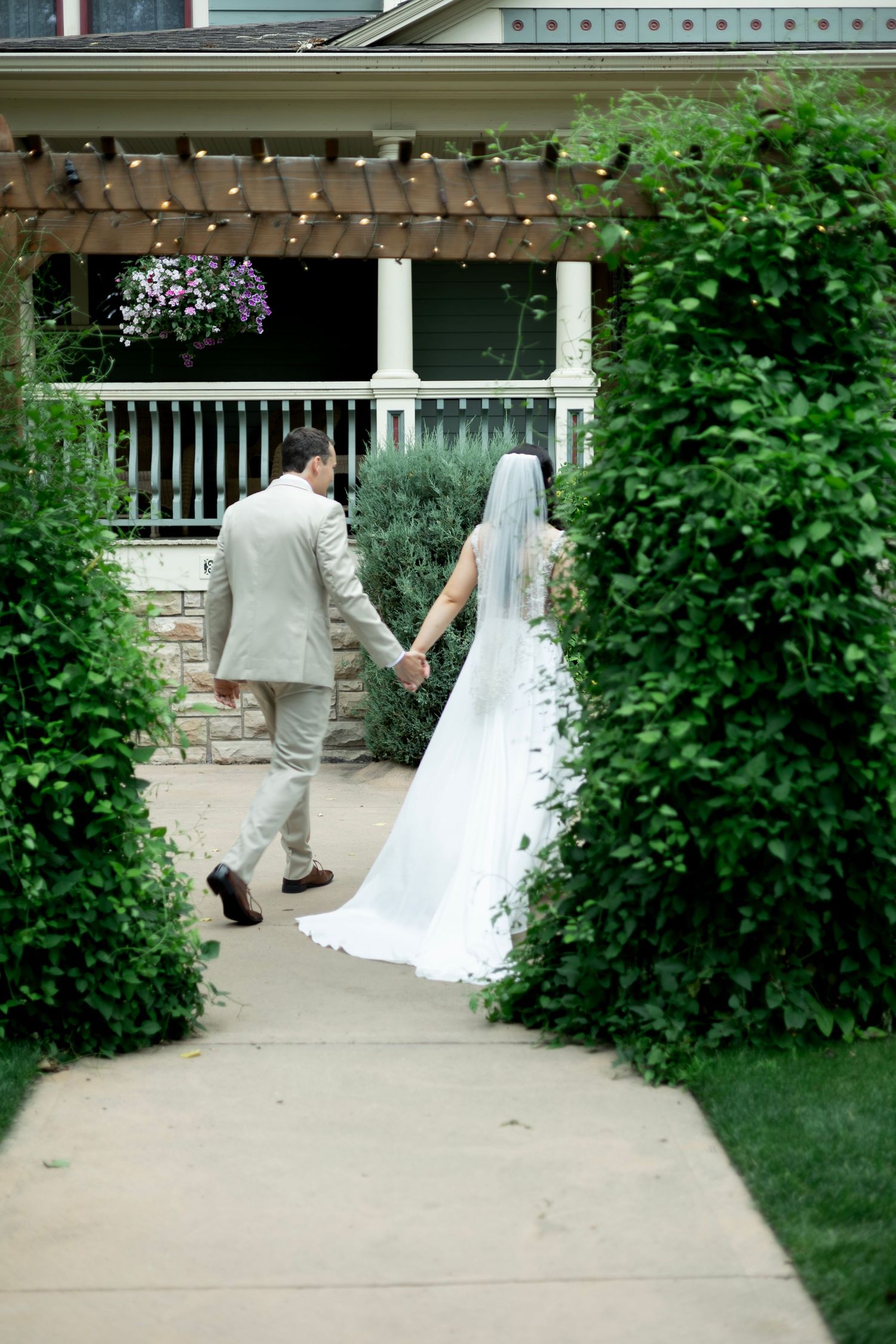 Bride in flowing white gown and groom in beige suit walk hand-in-hand through garden path towards covered porch.
