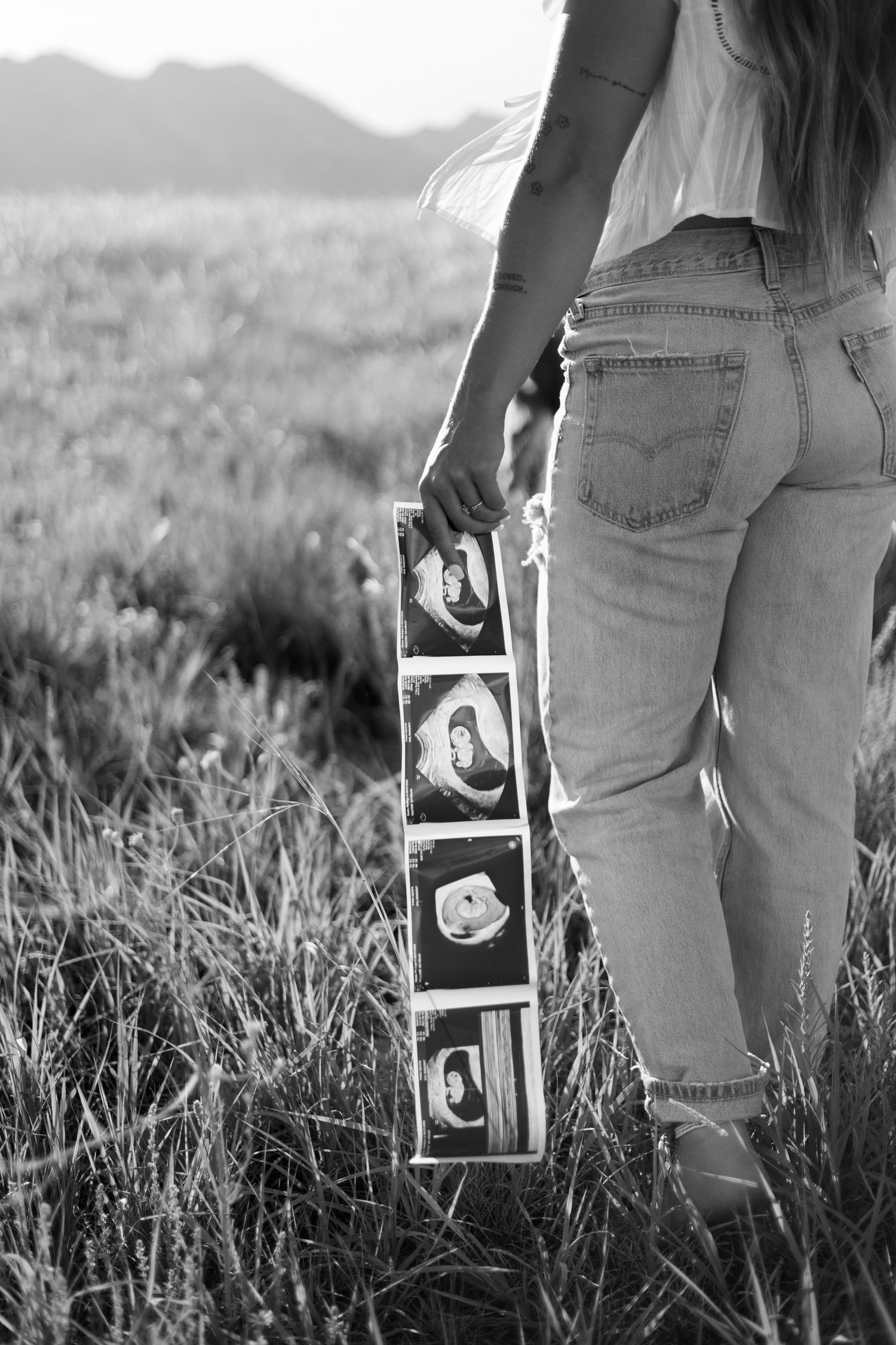 A black and white image showing someone holding ultrasound photos in tall grass with ripped jeans.