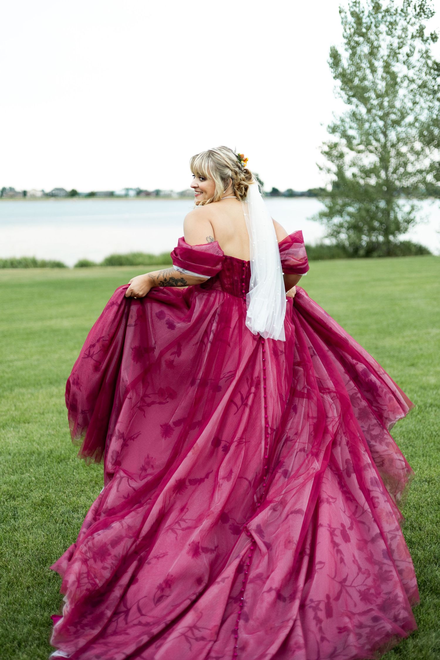 Bride in flowing burgundy ballgown twirls on lakeside lawn.