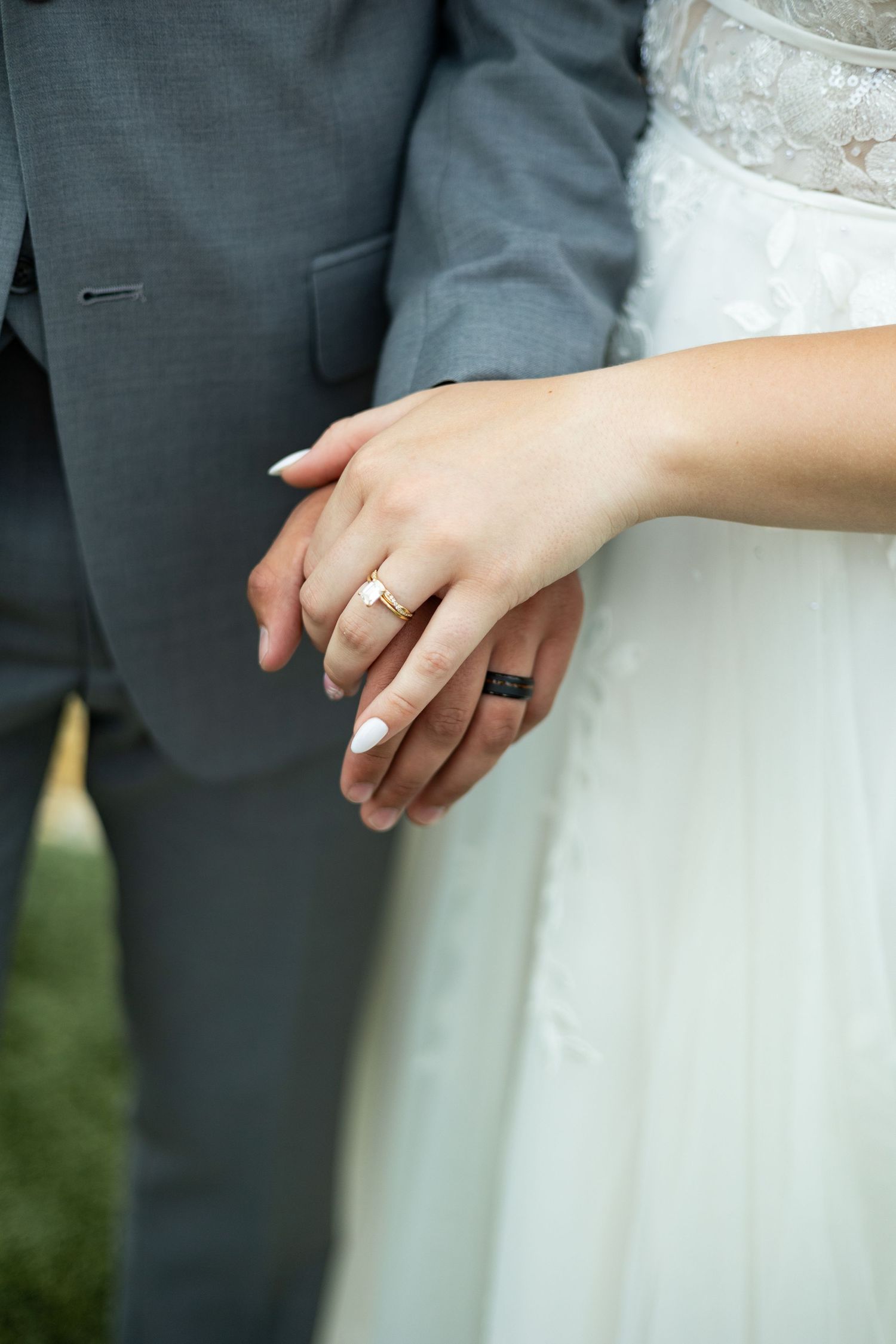 Close up of wedding rings and clasped hands showing a grey suit and white dress.