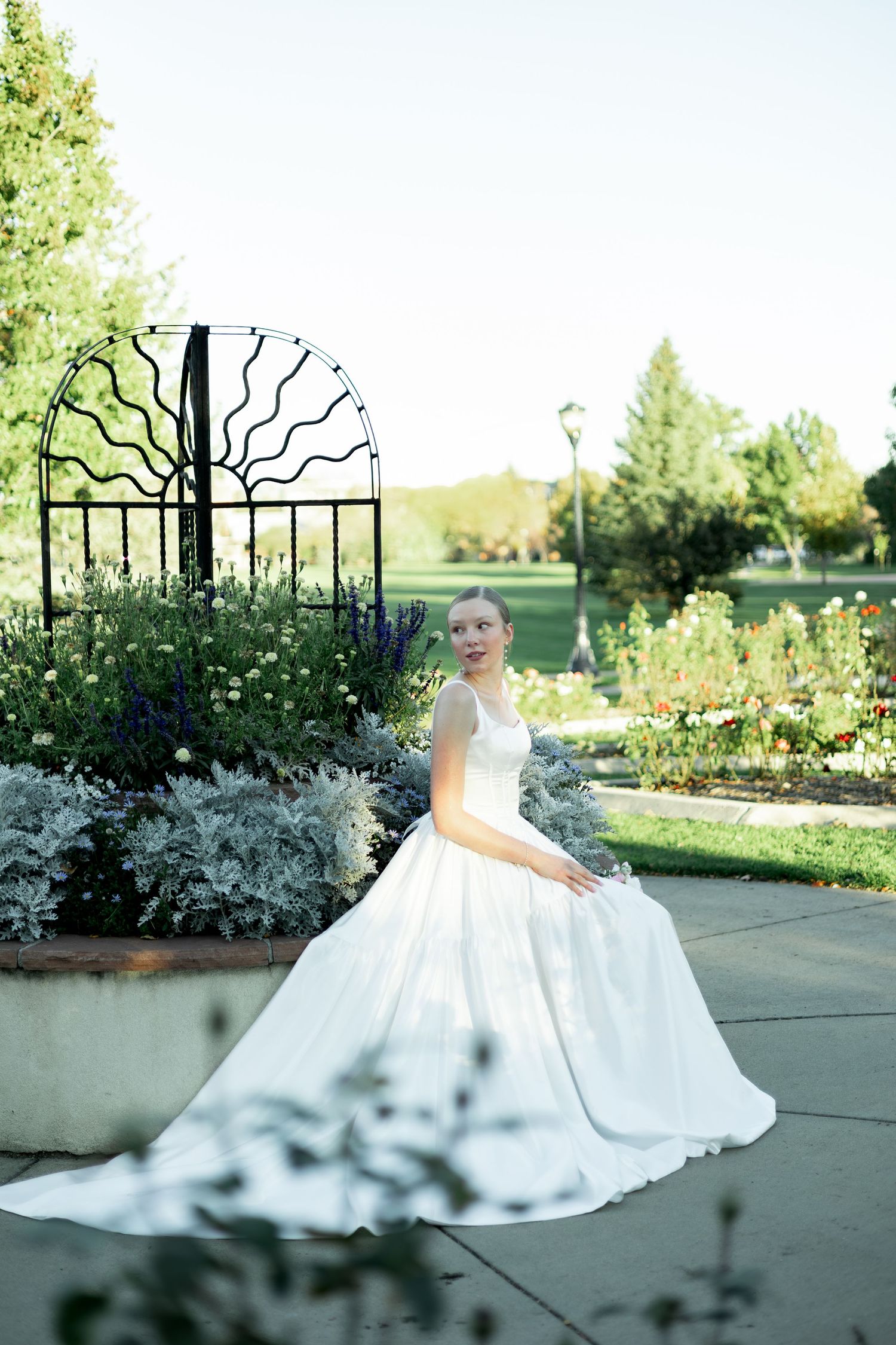 Bride in white ball gown sitting gracefully in a garden with colorful flowers and decorative iron gate backdrop.