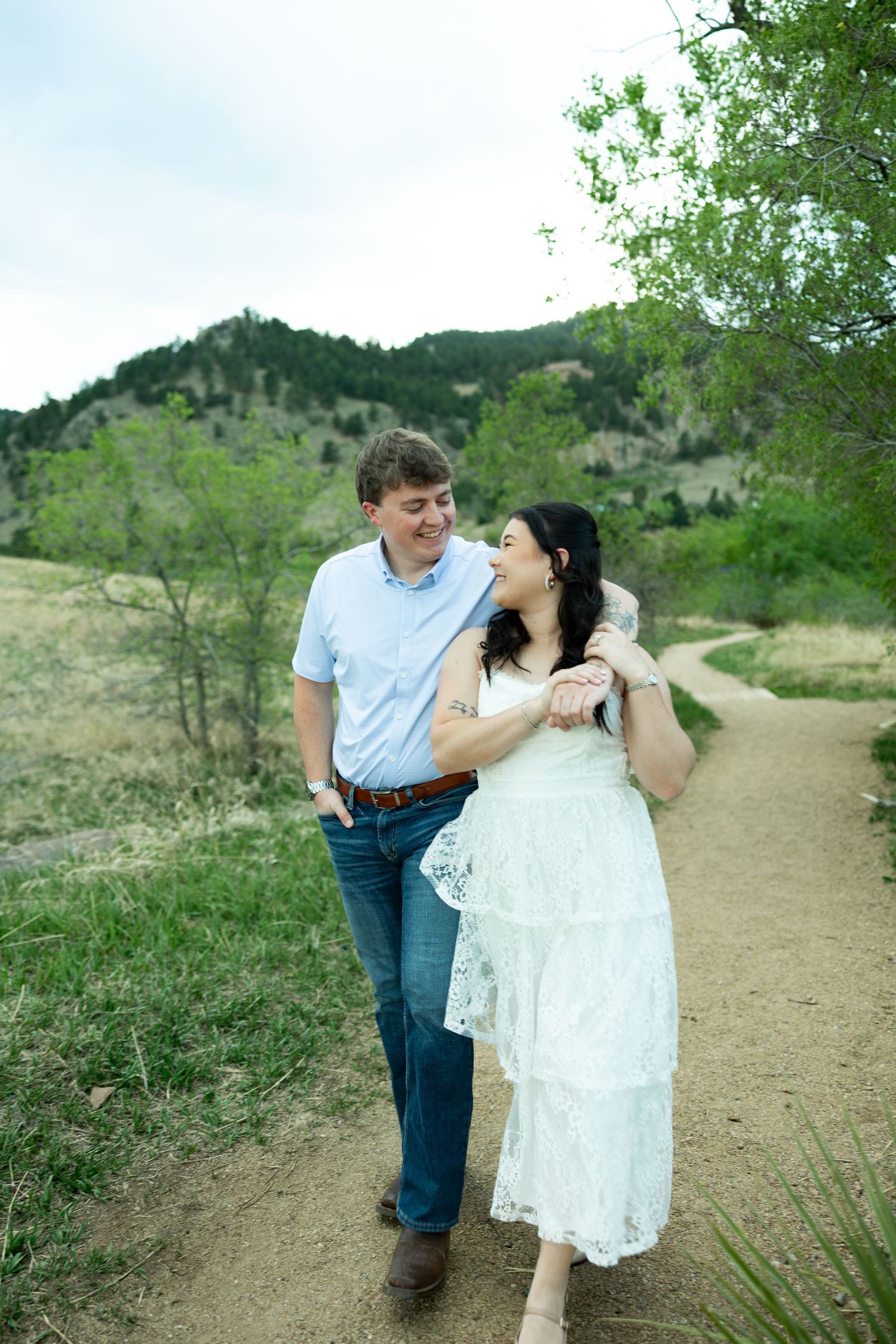 Happy couple walking on a dirt path through green Colorado foothills, woman in white lace dress embracing man in blue shirt.