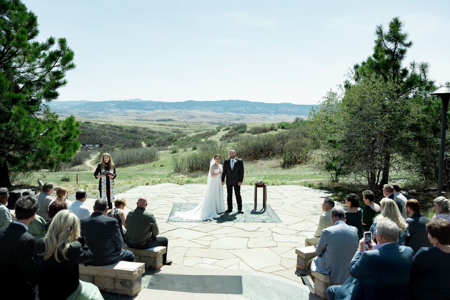 Outdoor wedding ceremony on stone patio with panoramic valley views, guests seated as couple stands at altar.