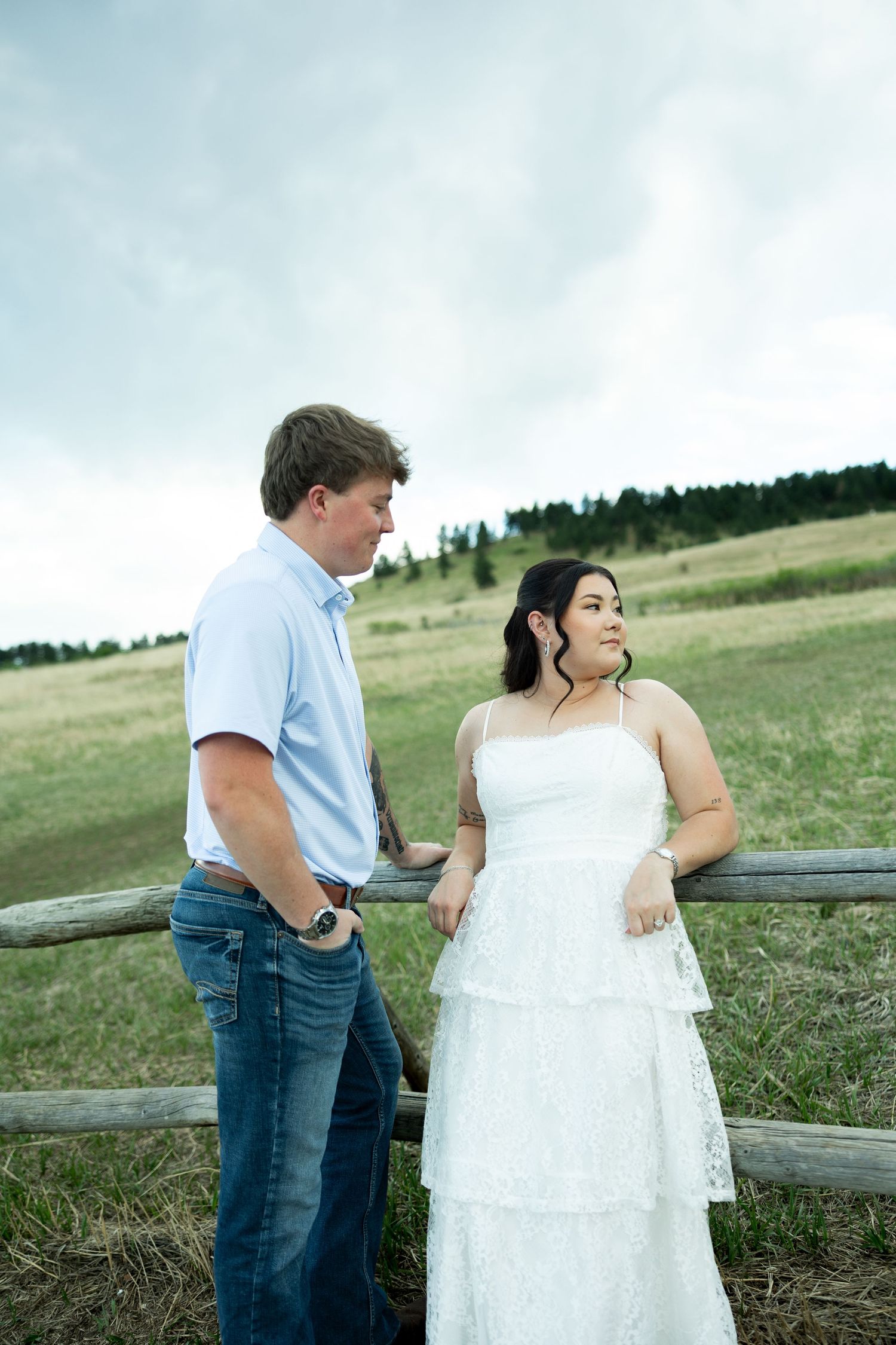 Couple in rustic field, man in blue shirt and jeans, woman in white lace dress, leaning on wooden fence.