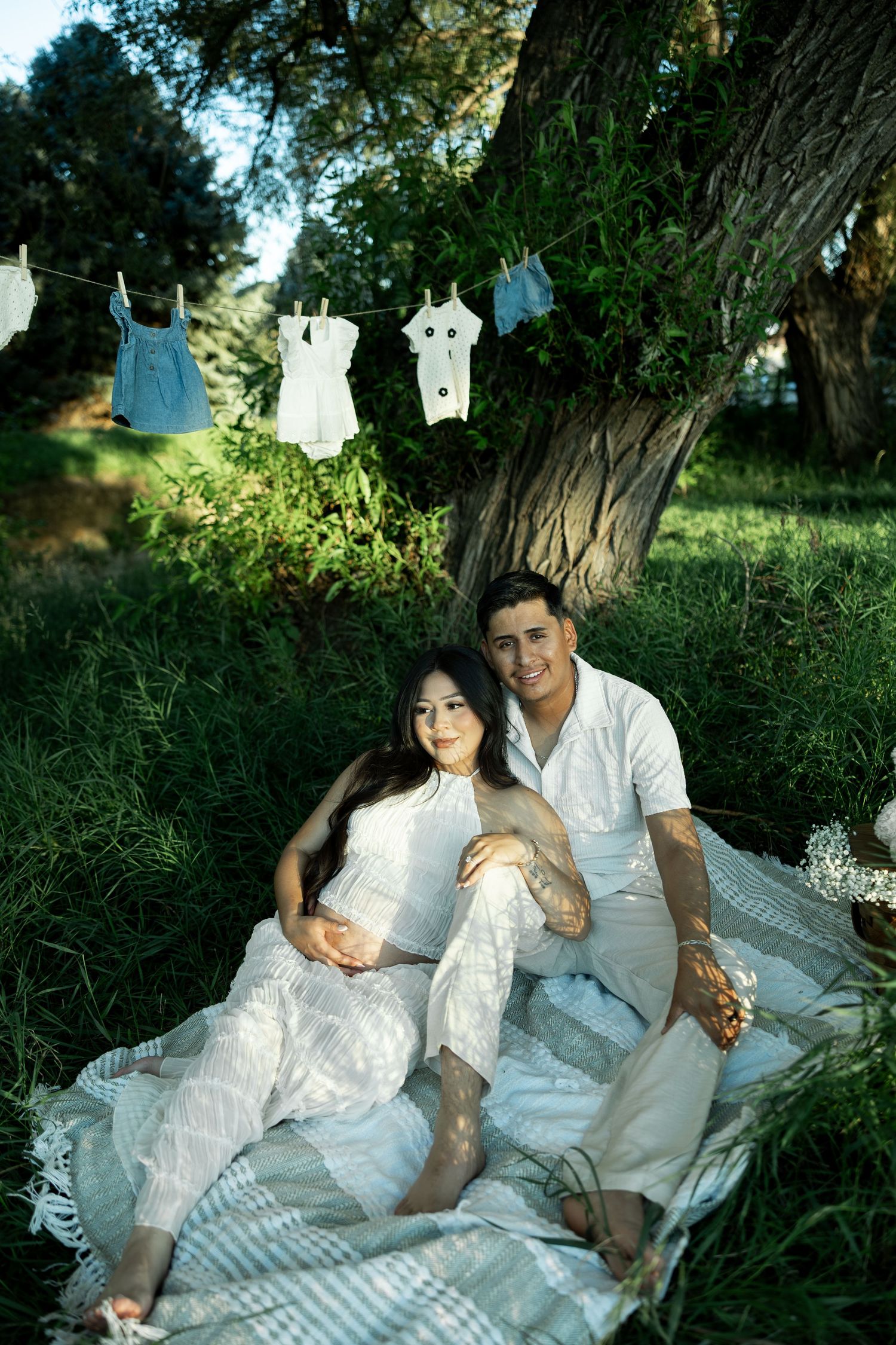 Pregnant couple in white outfits sitting under a tree with baby clothes hanging on a line in a lush green park.
