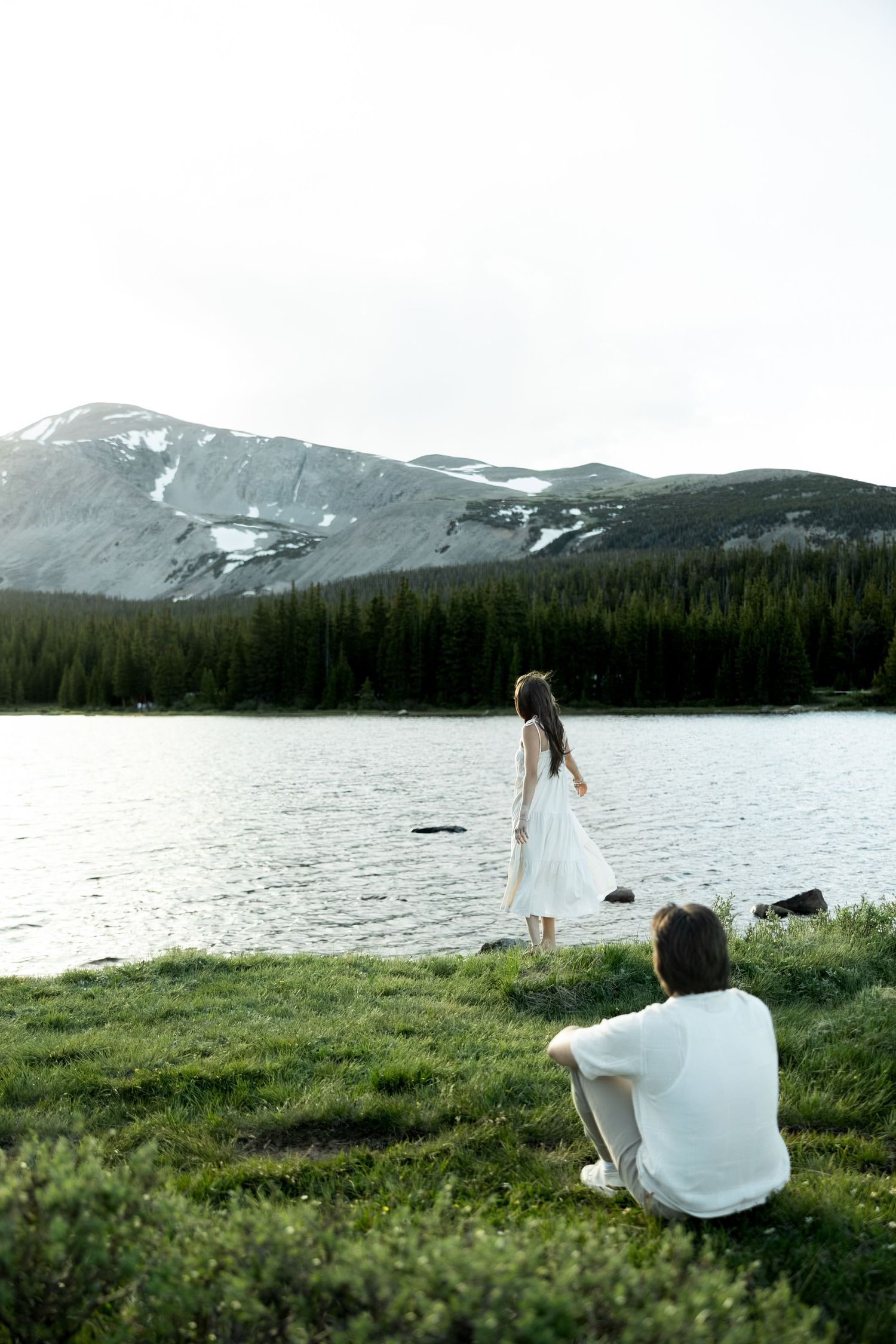 Couple in white by a mountain lake, woman standing at water's edge, man sitting on grass, snow-capped peaks behind.