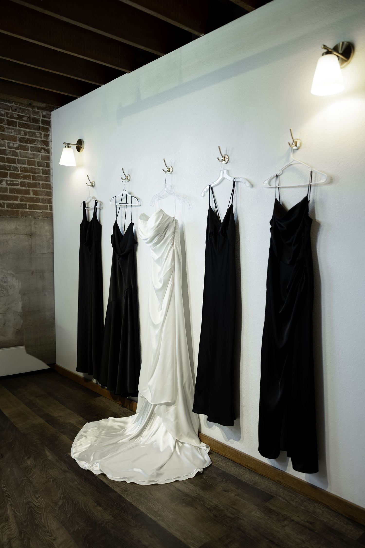 White wedding gown with train hangs among black bridesmaid dresses on wall hooks in rustic venue with brick walls.