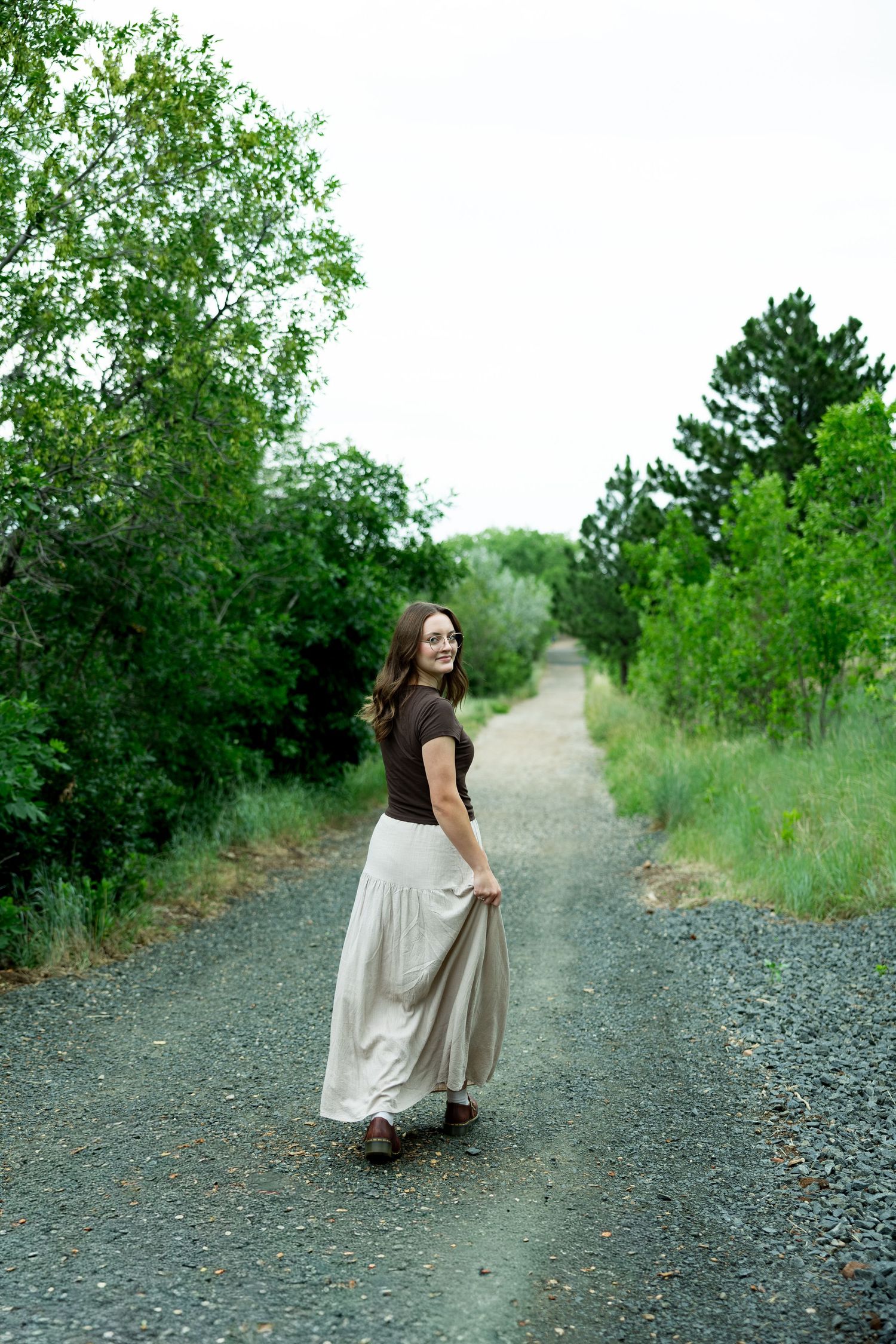 Young woman in brown top and flowing beige skirt smiling on a gravel path surrounded by lush green trees.