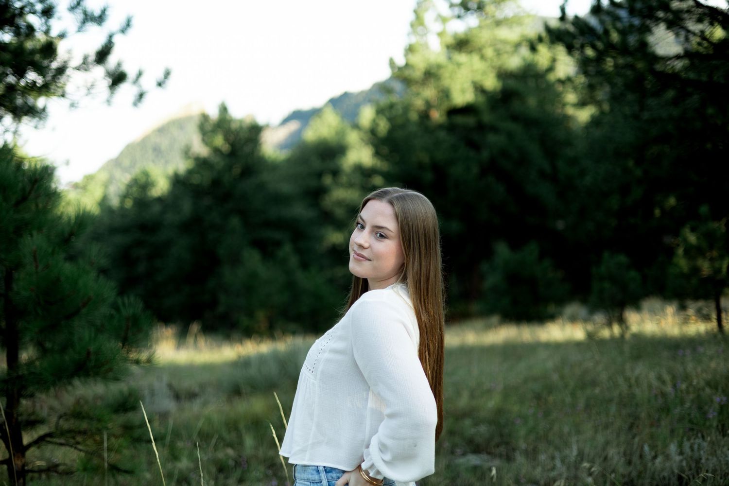 A woman in a white dress sits thoughtfully in a sunlit meadow with mountains and trees in the background.