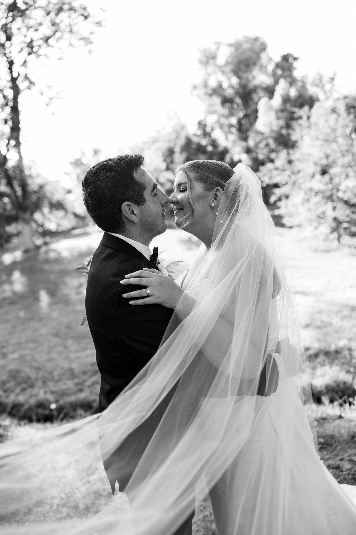 Black and white photo of a bride and groom sharing a joyful moment, her veil flowing, surrounded by lush trees.