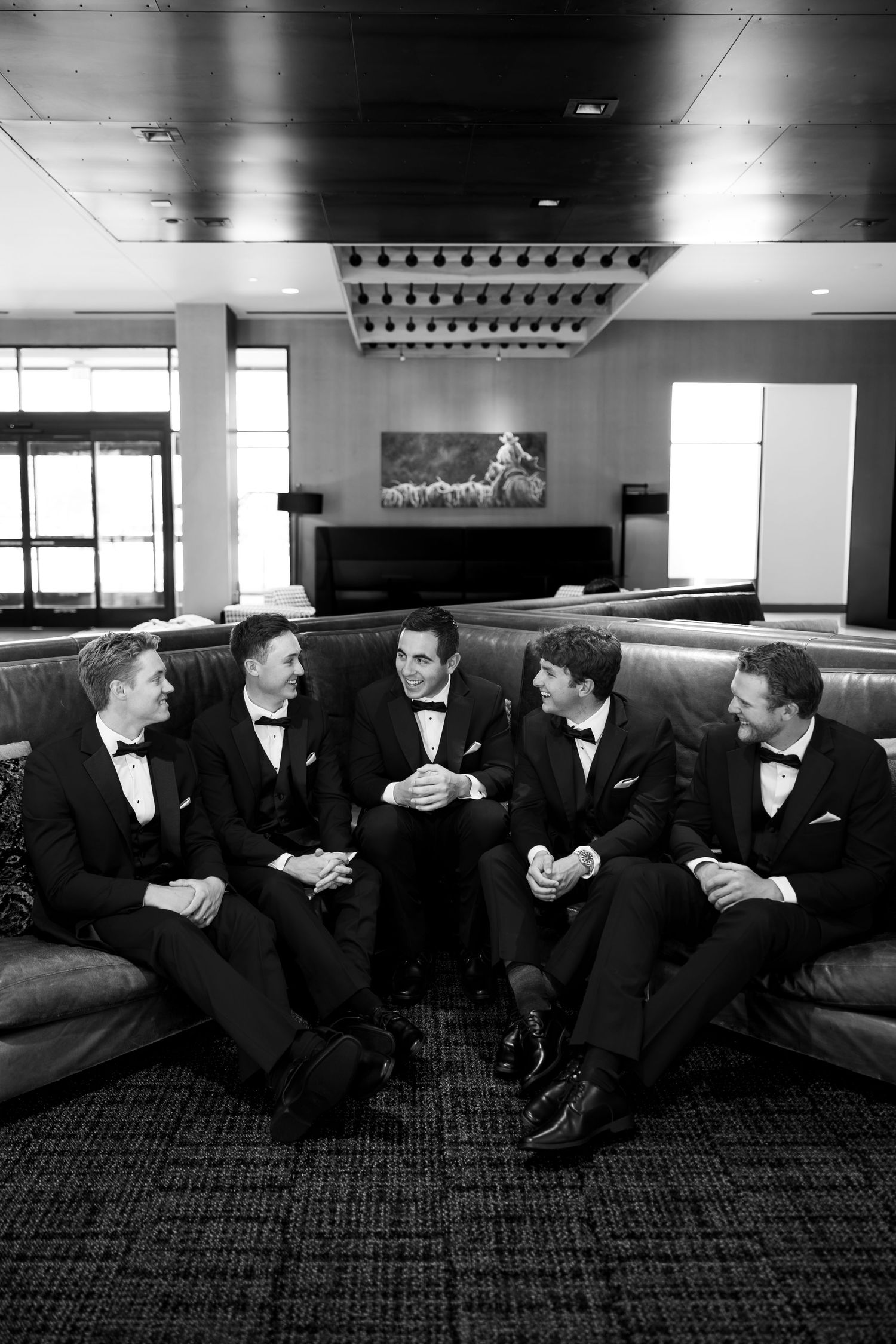 Five groomsmen in black tuxedos laughing together on a couch in an elegant venue, black and white photo.