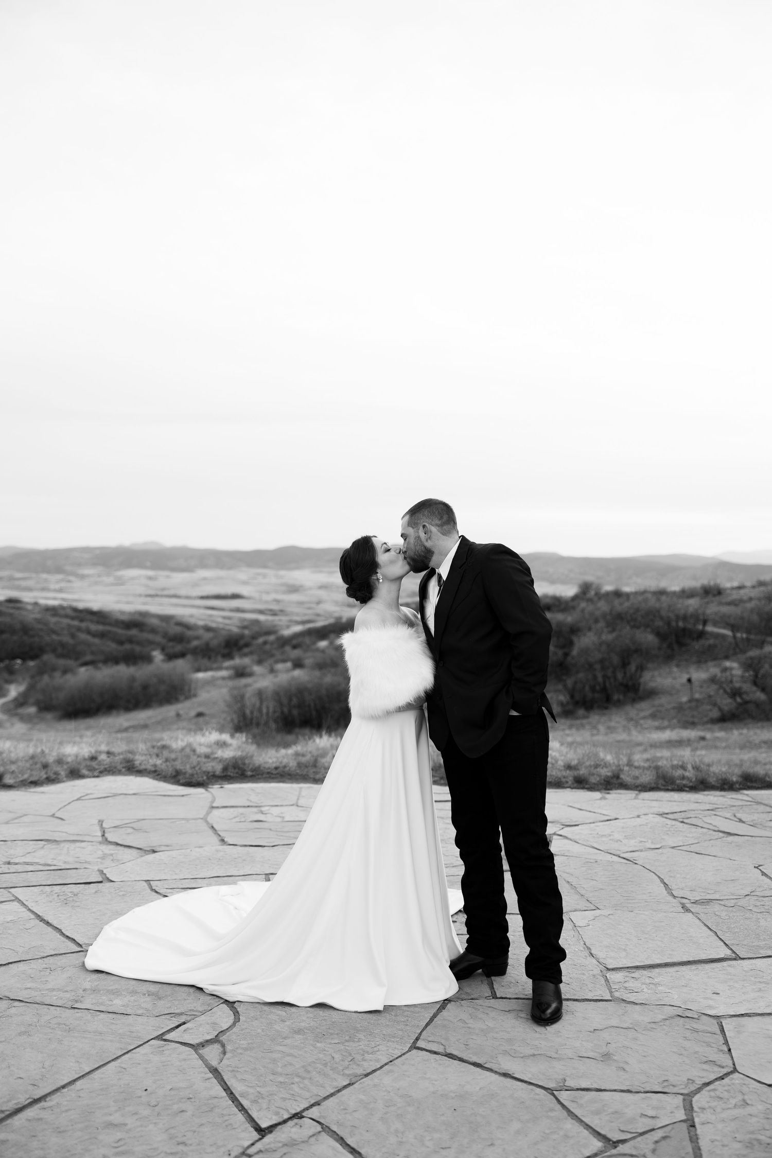 Black and white photo of a bride and groom sharing a kiss on a stone terrace with scenic rolling hills in the background.
