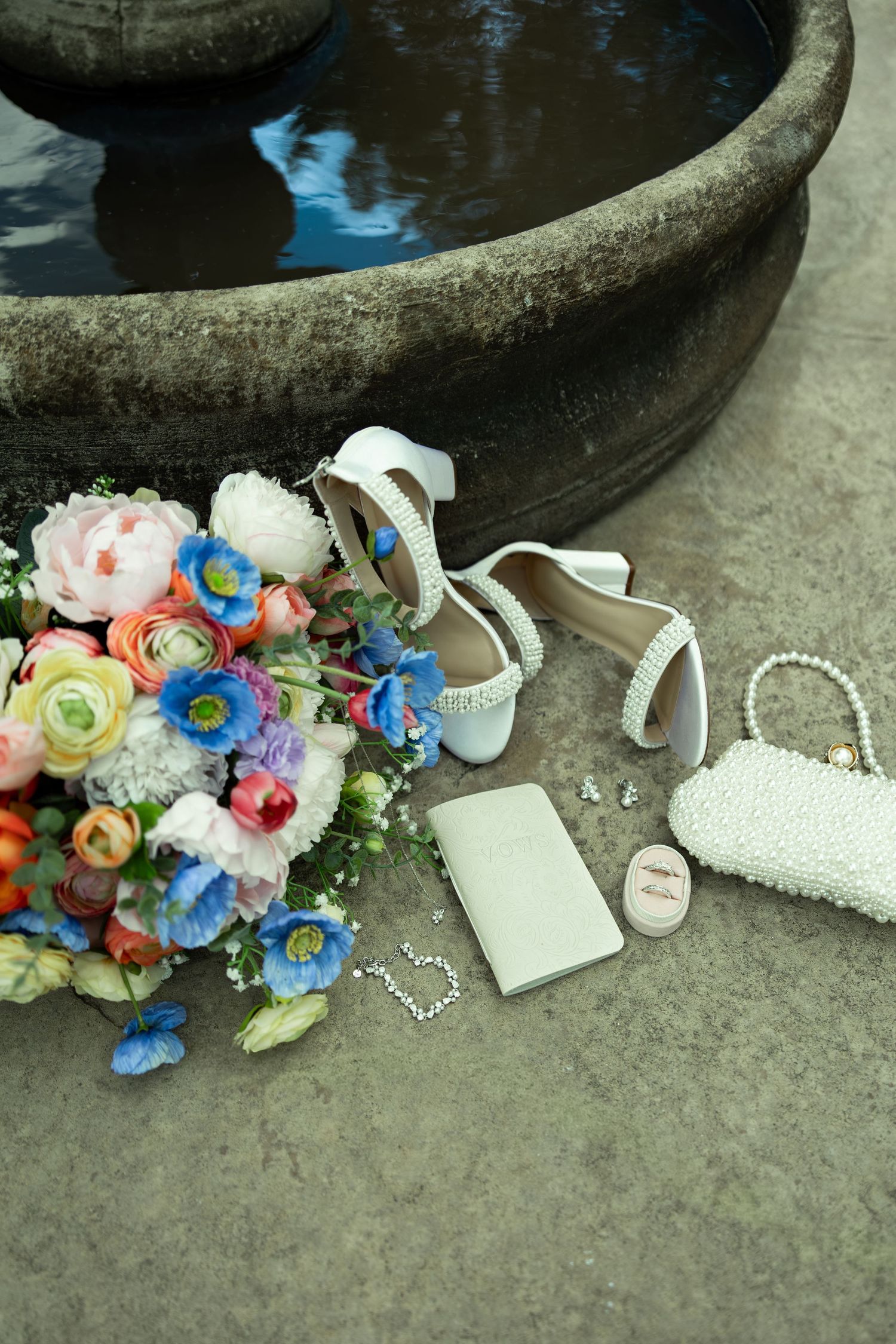 Bridal accessories arranged near stone fountain, featuring white heels, pearl clutch, bouquet of colorful flowers, and jewelry.