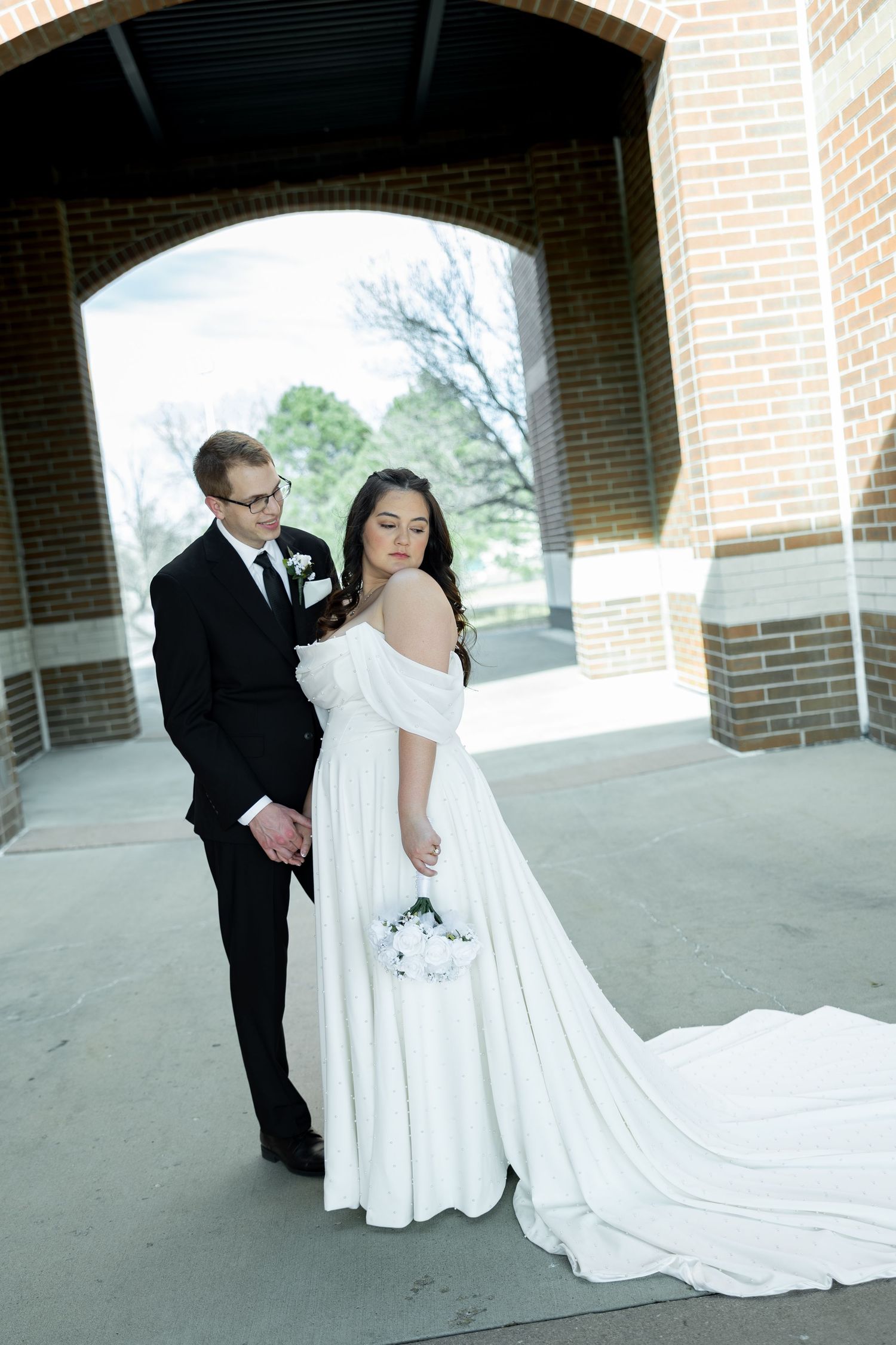 Bride in white off-shoulder gown and groom in black tuxedo pose together under brick archway for wedding portrait.