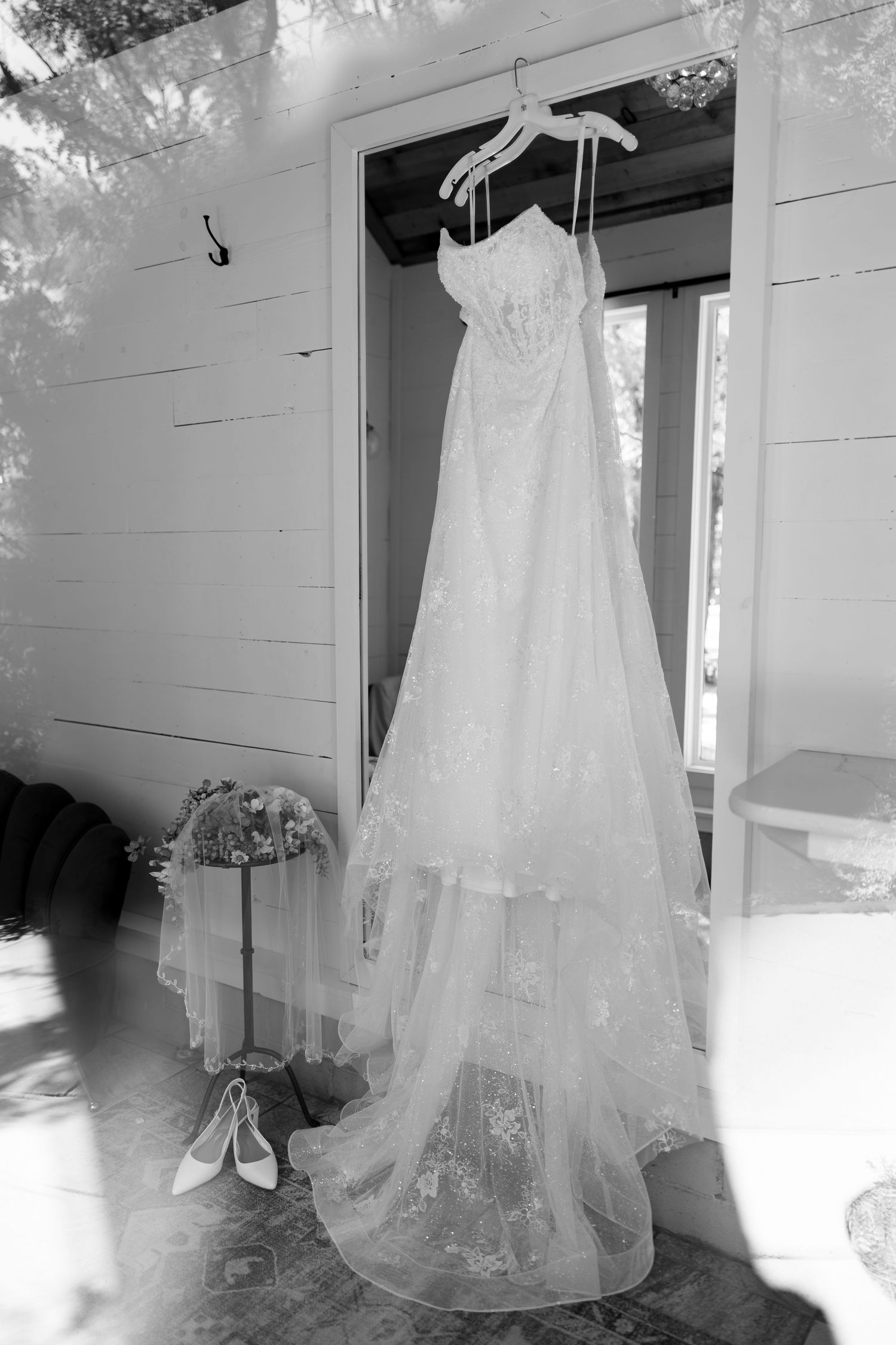 Black and white photo of an elegant lace wedding dress hanging in a doorway with bridal shoes and bouquet nearby.