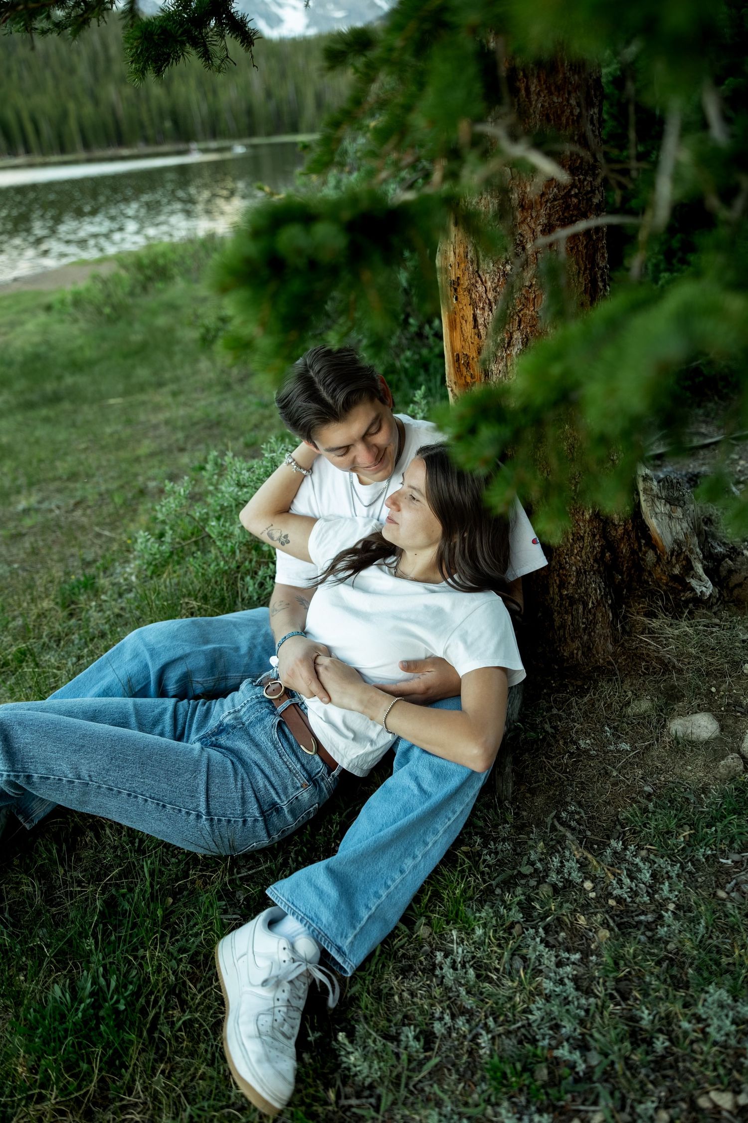 Couple relaxing under a pine tree by a mountain lake, woman leaning against man in white tees.