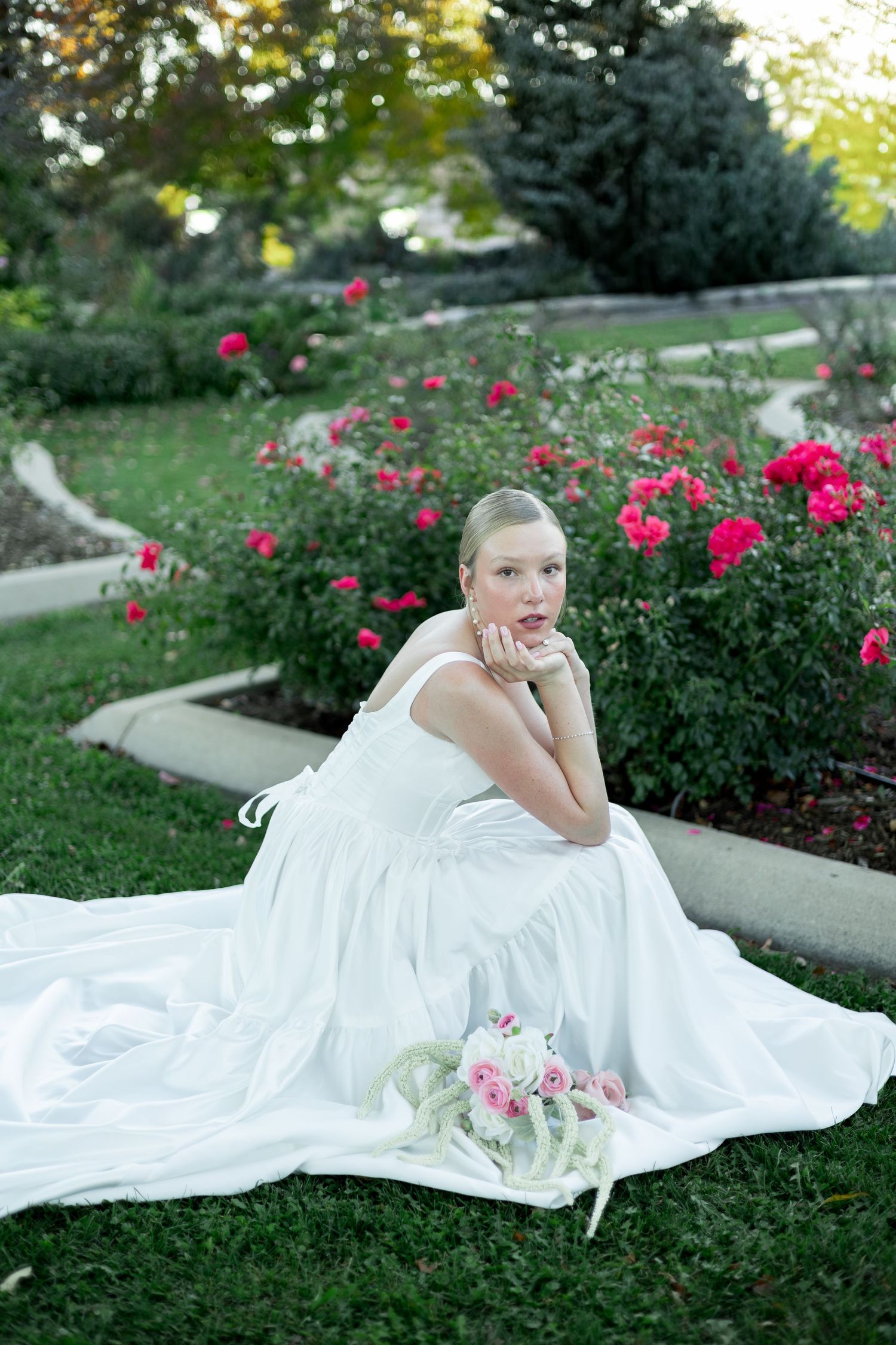 Bride in white gown sitting on grass beside pink roses garden, holding bouquet, looking elegant.