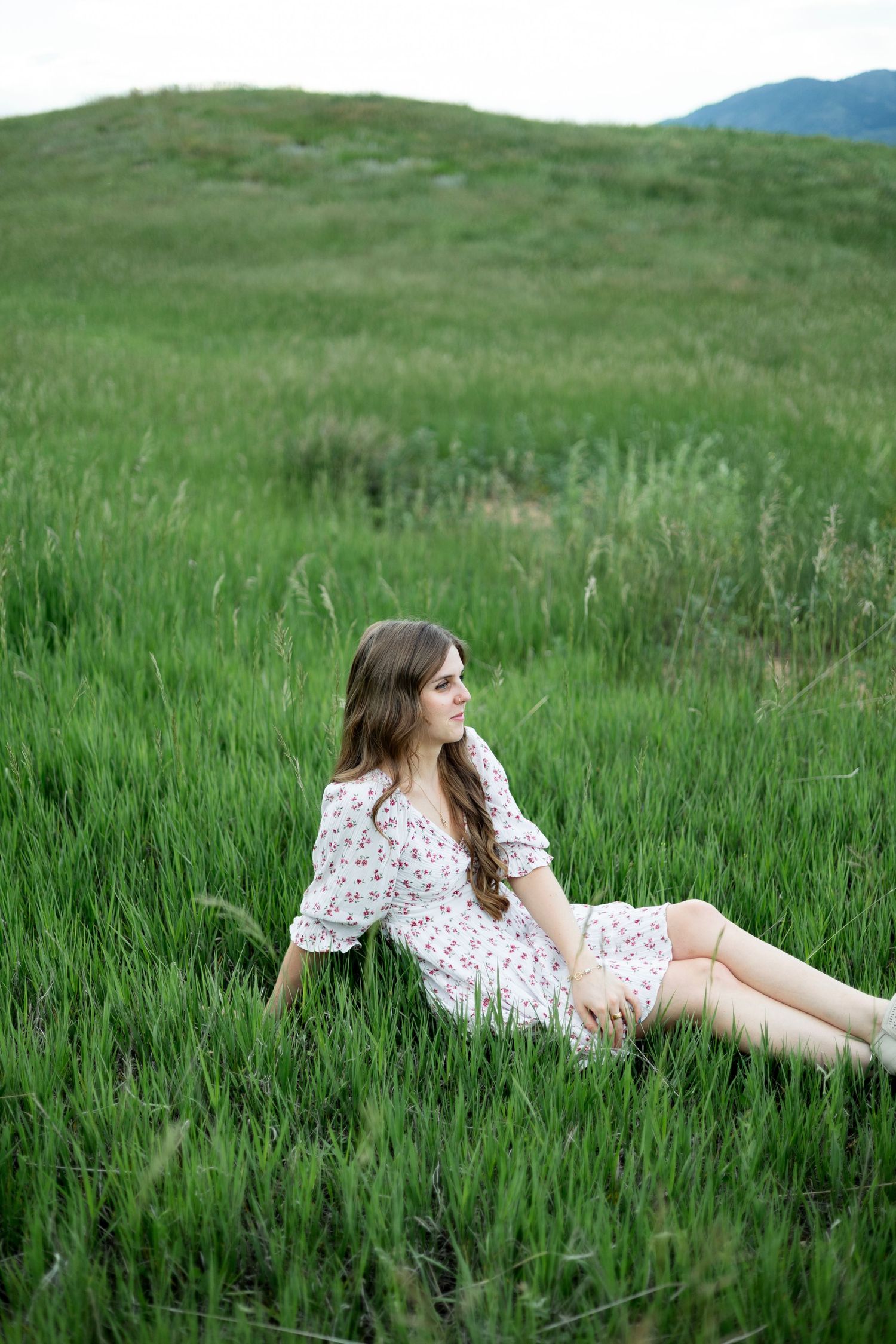 Young woman in floral dress sitting in lush green meadow with rolling hills in background.