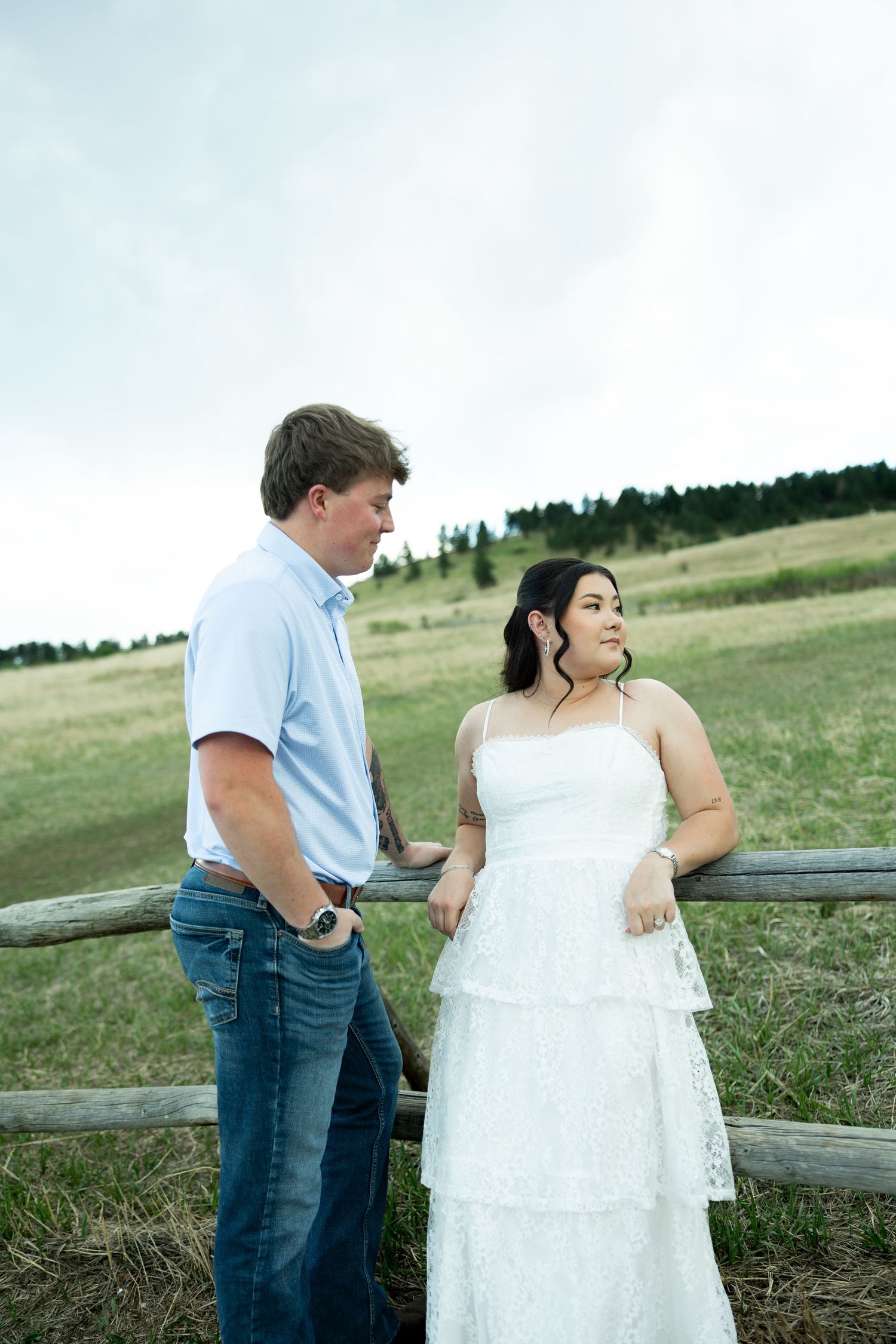 Bride in white dress and groom in blue shirt share a tender moment leaning against a rustic wooden fence outdoors.