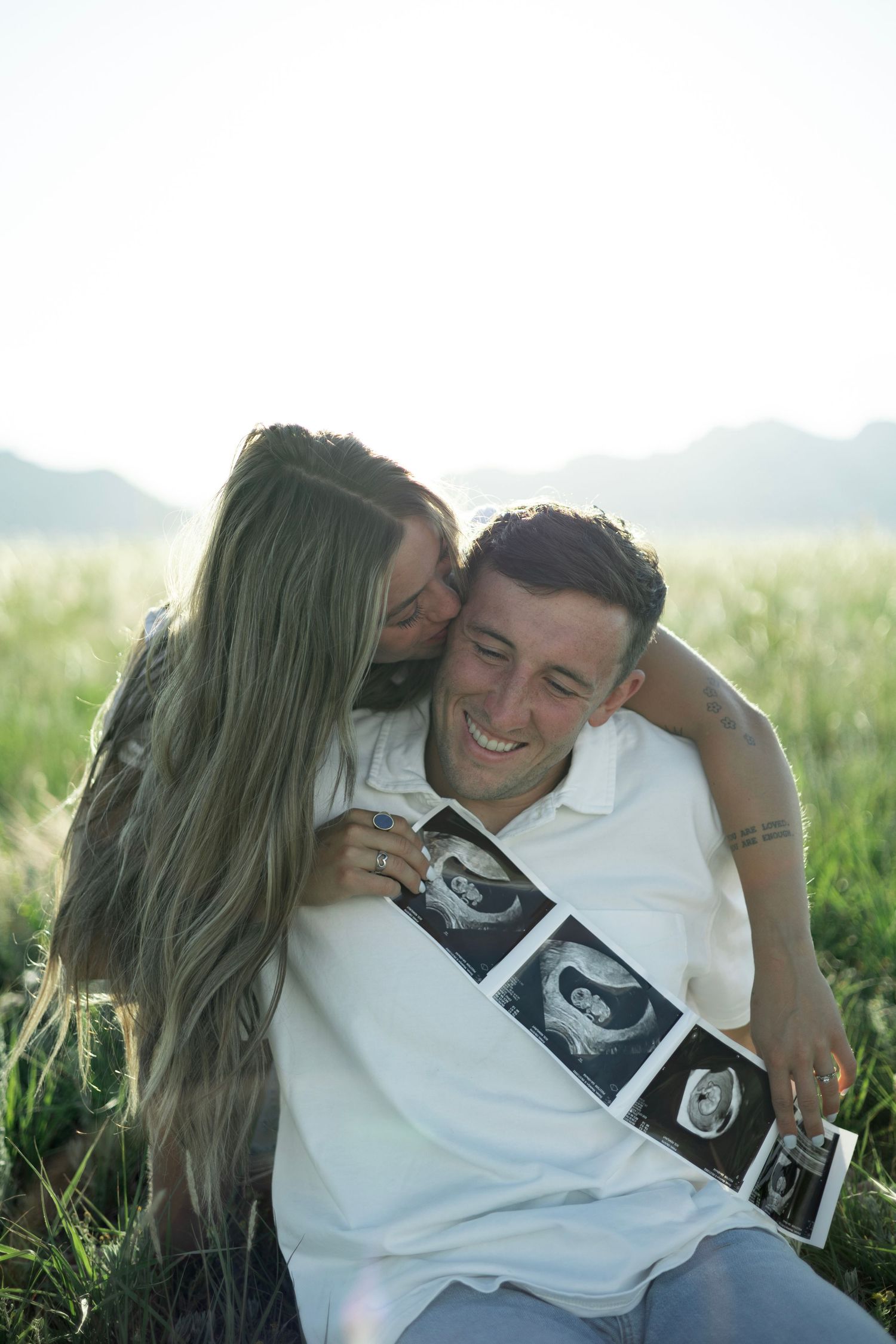 A couple sits in a grassy field, sharing a tender moment while looking at ultrasound photos together.