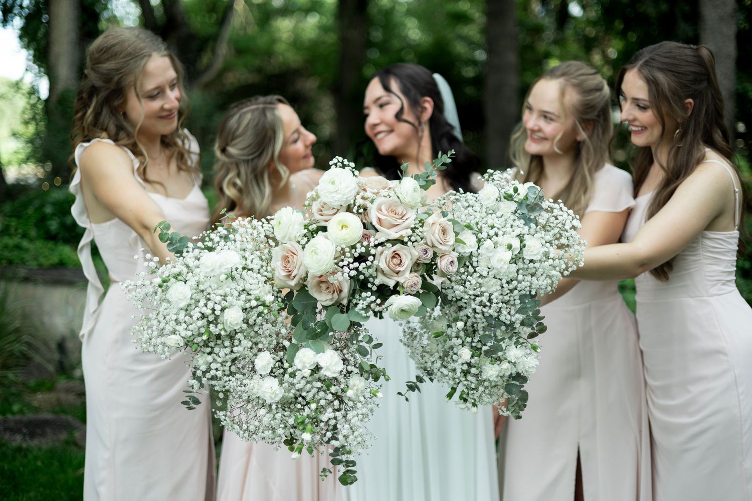 Bridesmaids in light dresses gather around stunning white floral bouquet during outdoor wedding celebration.