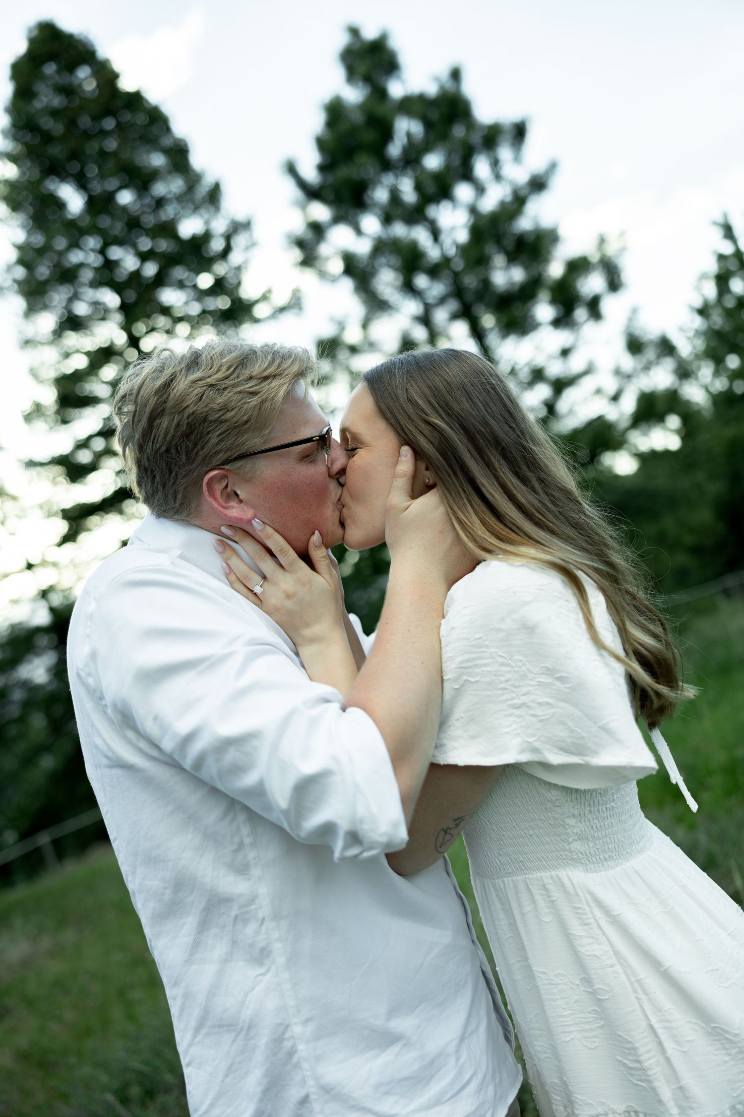 Couple sharing a tender kiss outdoors with tall green pine trees in the background.