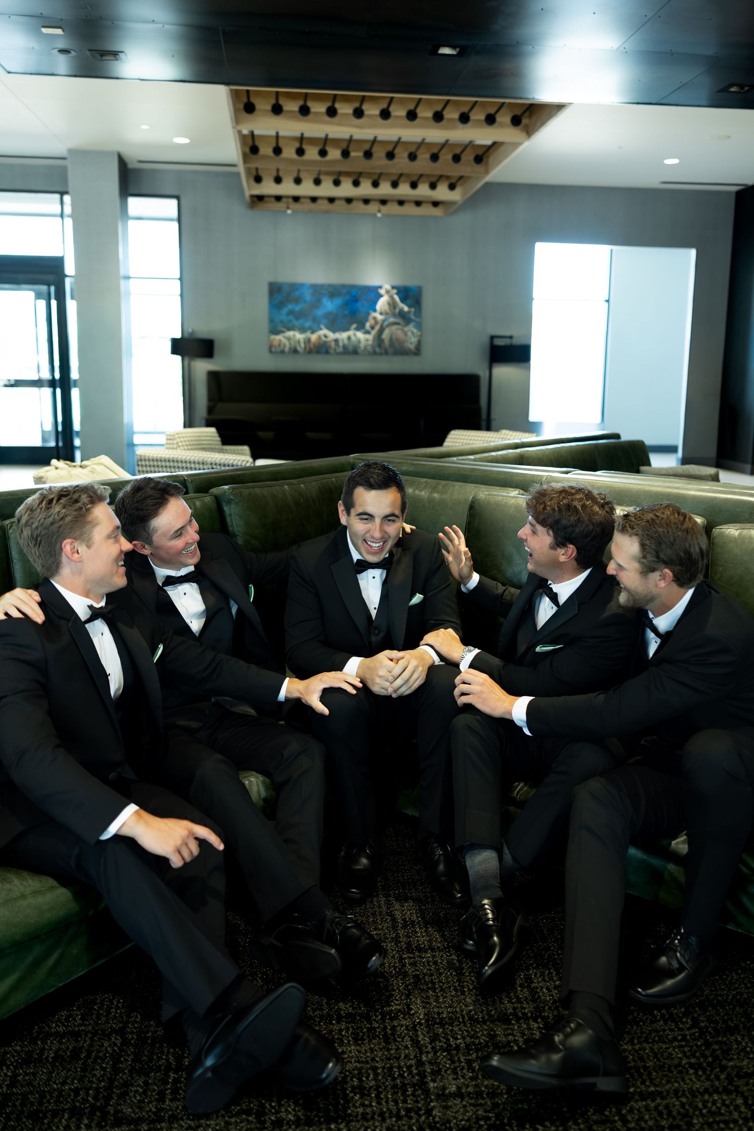Five groomsmen in black tuxedos laugh together on a green sofa in an elegant hotel lobby.