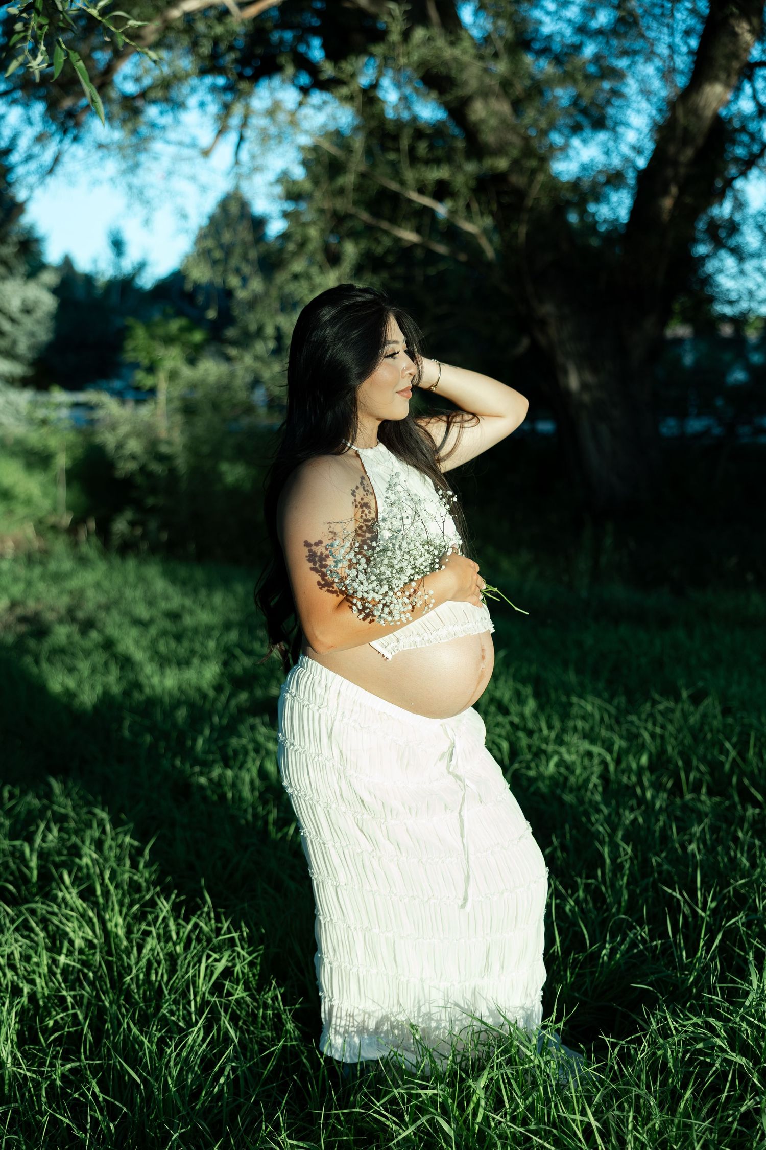 Pregnant woman in white dress holding wildflowers stands in lush green grass under trees in soft natural sunlight.