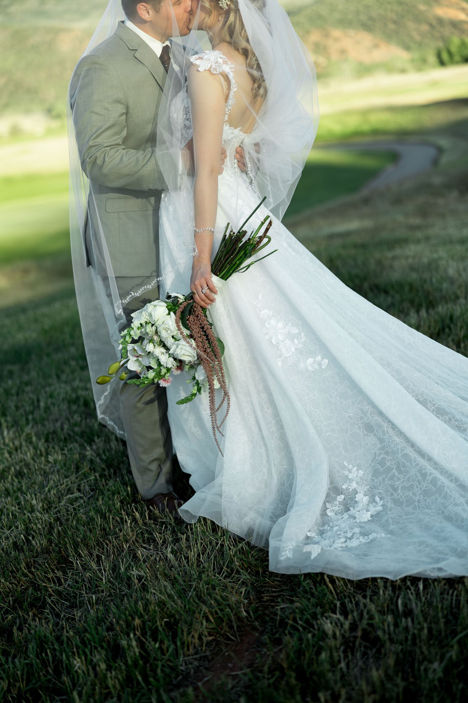 Bride and groom share a kiss outdoors, bride in lace gown with veil holding white bouquet on green grass.