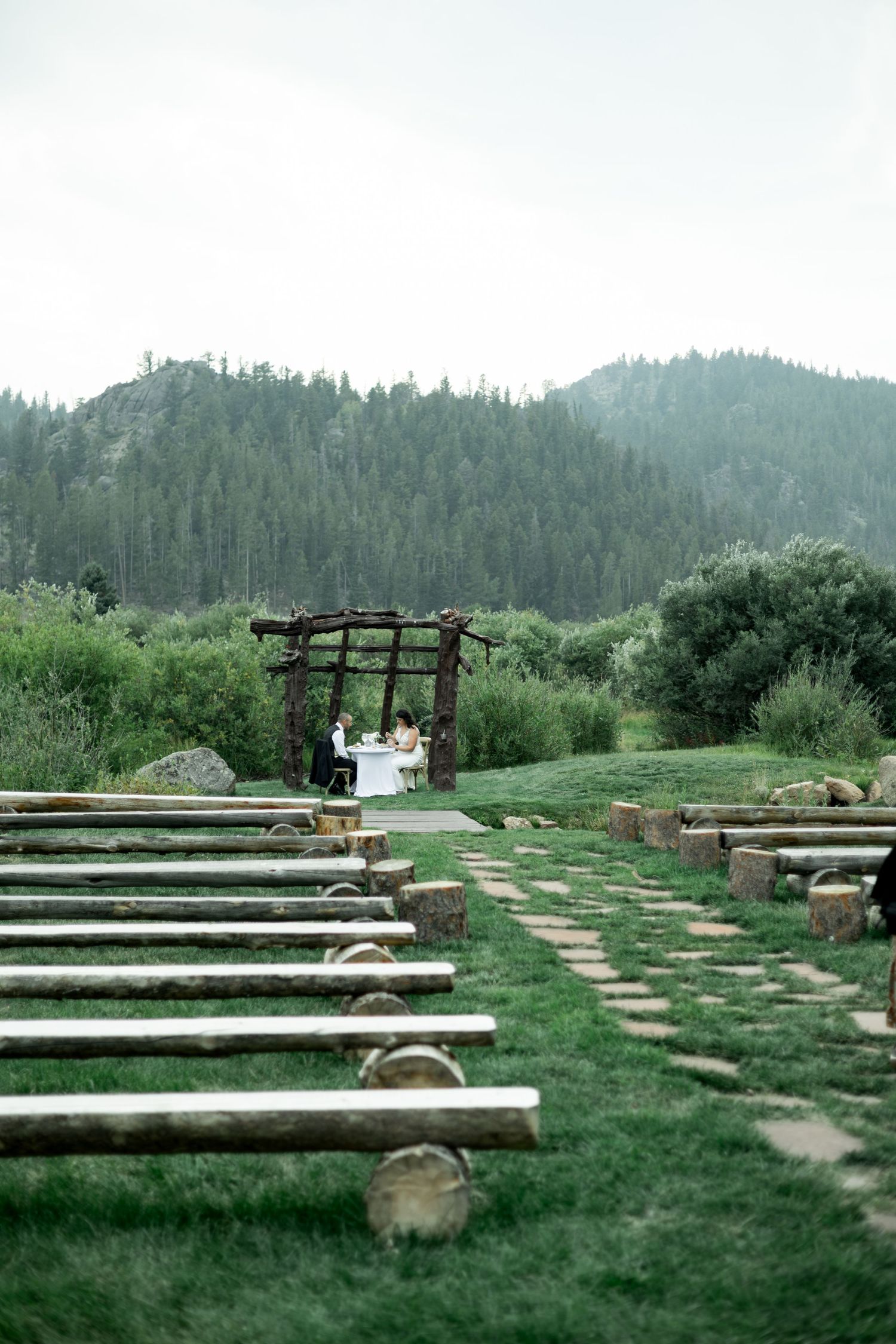 Outdoor wedding ceremony setup with wooden benches arranged in rows on a grassy hillside surrounded by forest.