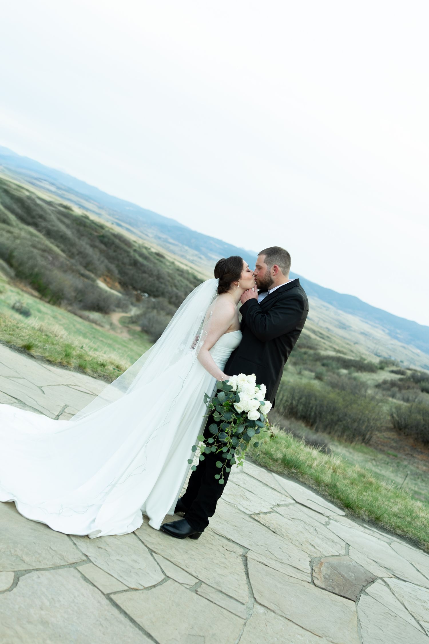 Bride and groom sharing a kiss on a stone terrace with scenic rolling hills in the background on their wedding day.