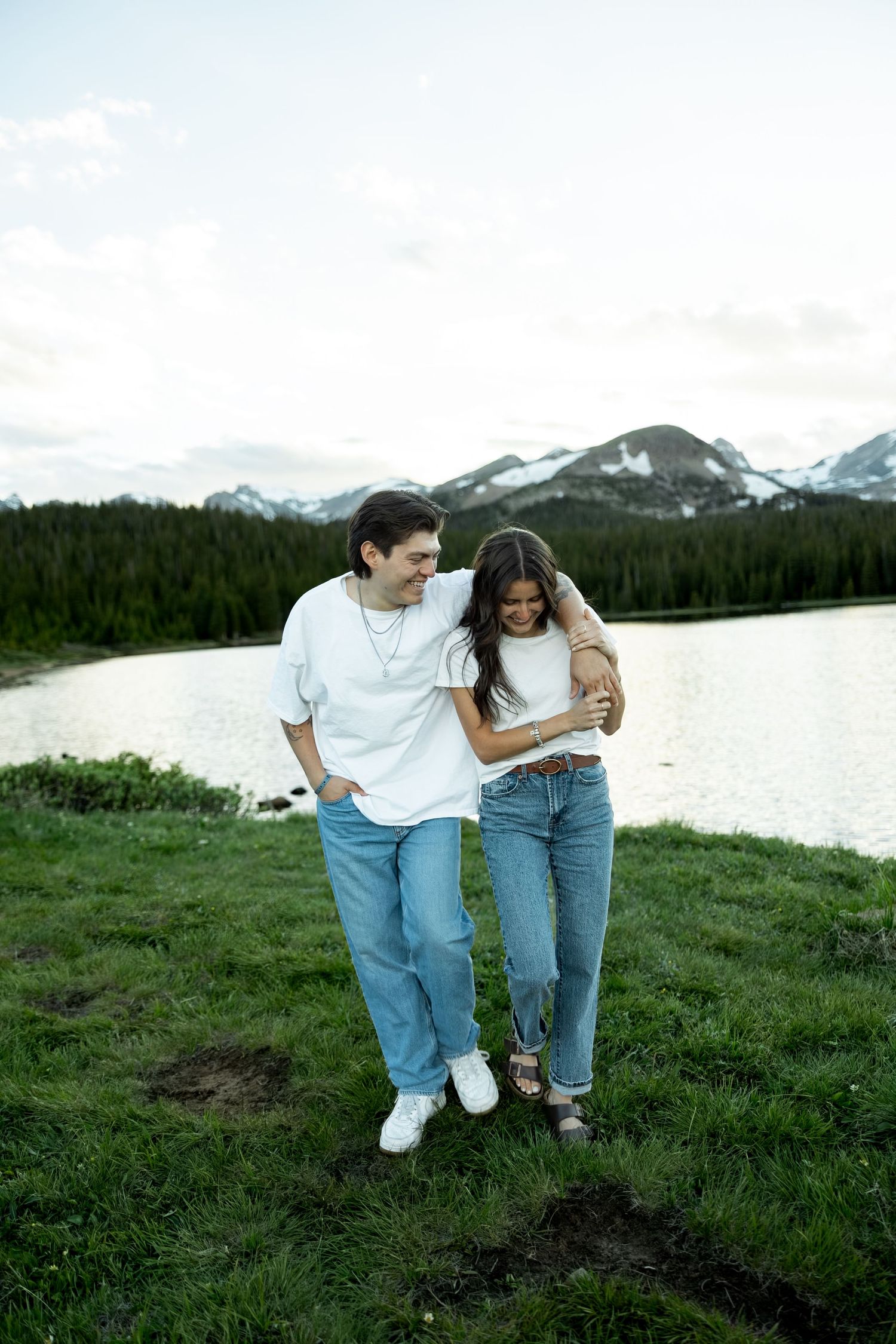 Couple in matching white tees and jeans embracing by a mountain lake with snow-capped peaks and pine forest backdrop.