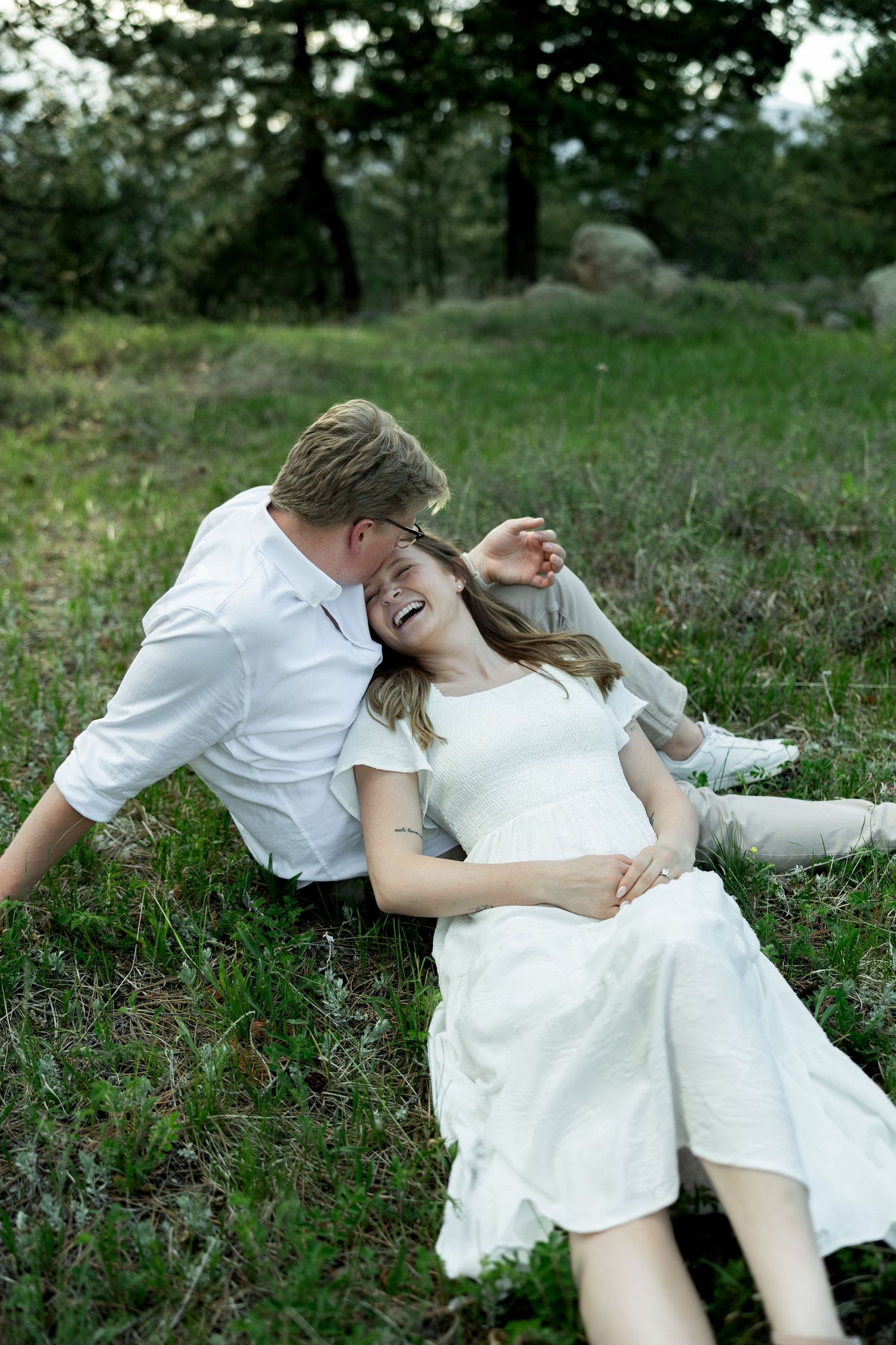 Happy couple laughing and relaxing on green grass in a forest, woman in white dress leaning back on man in white shirt.