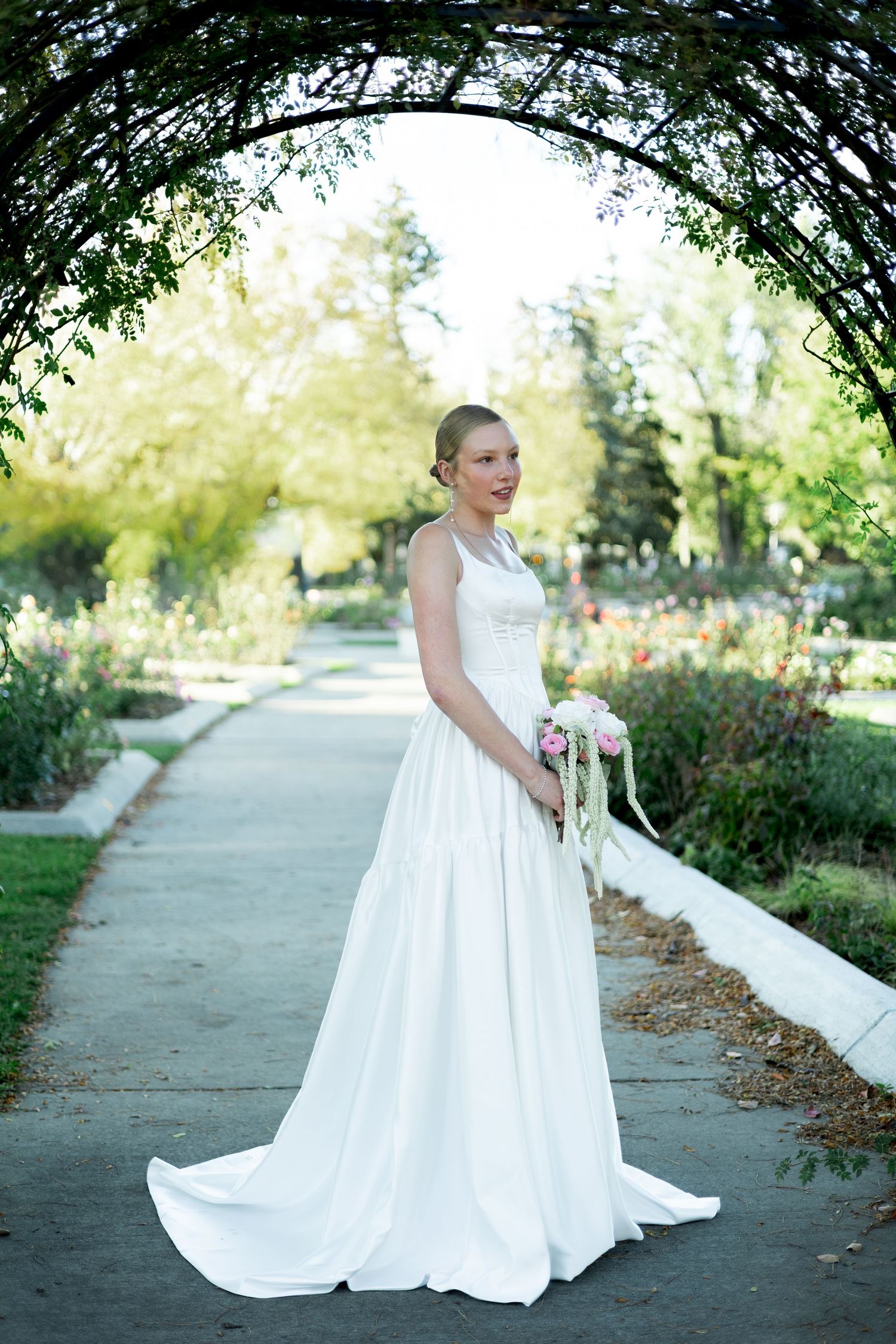 Bride in white gown holding pink bouquet stands under green arch in garden pathway.