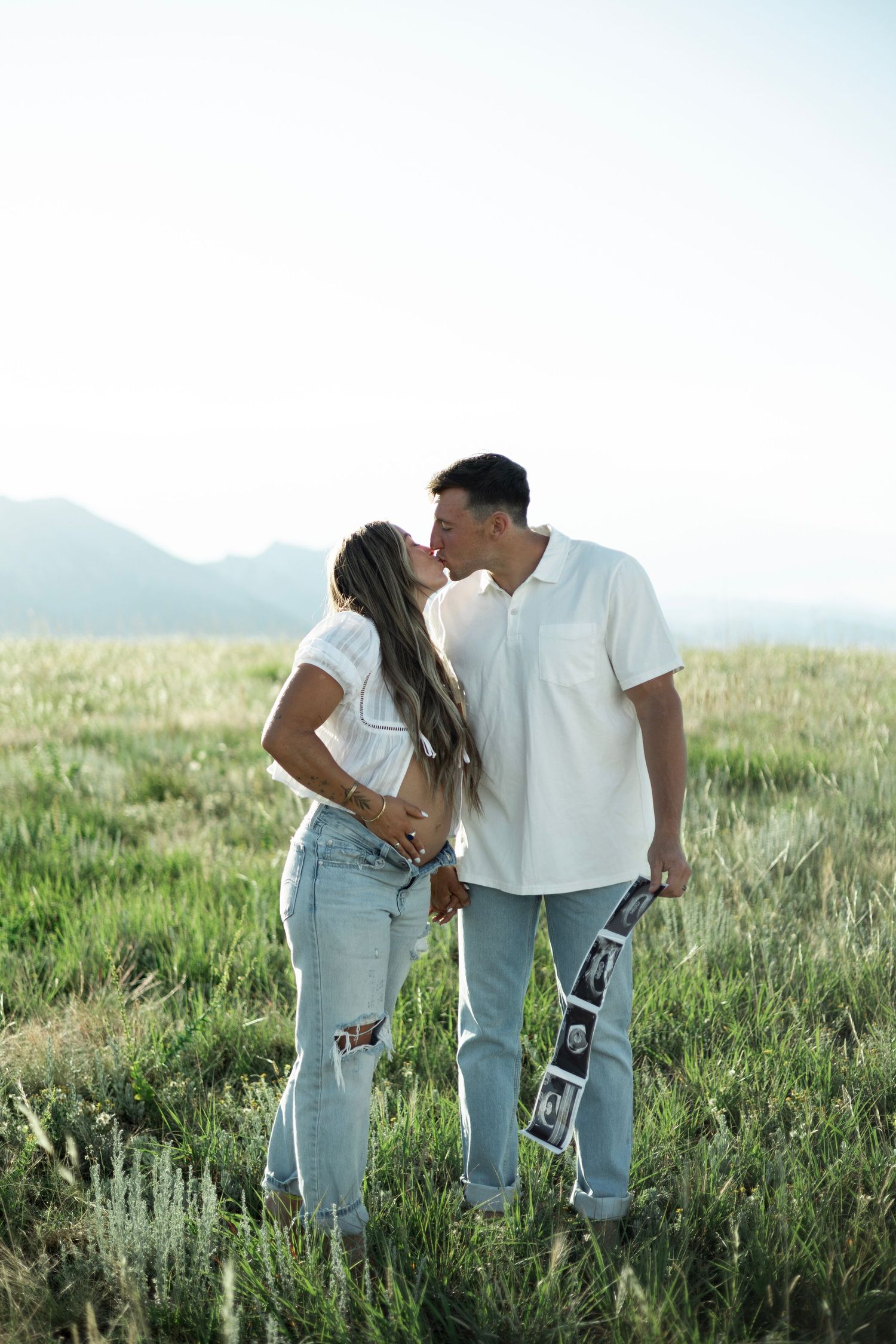 A couple in casual white tops and ripped jeans share a romantic moment while standing in a grassy meadow with mountains behind them.